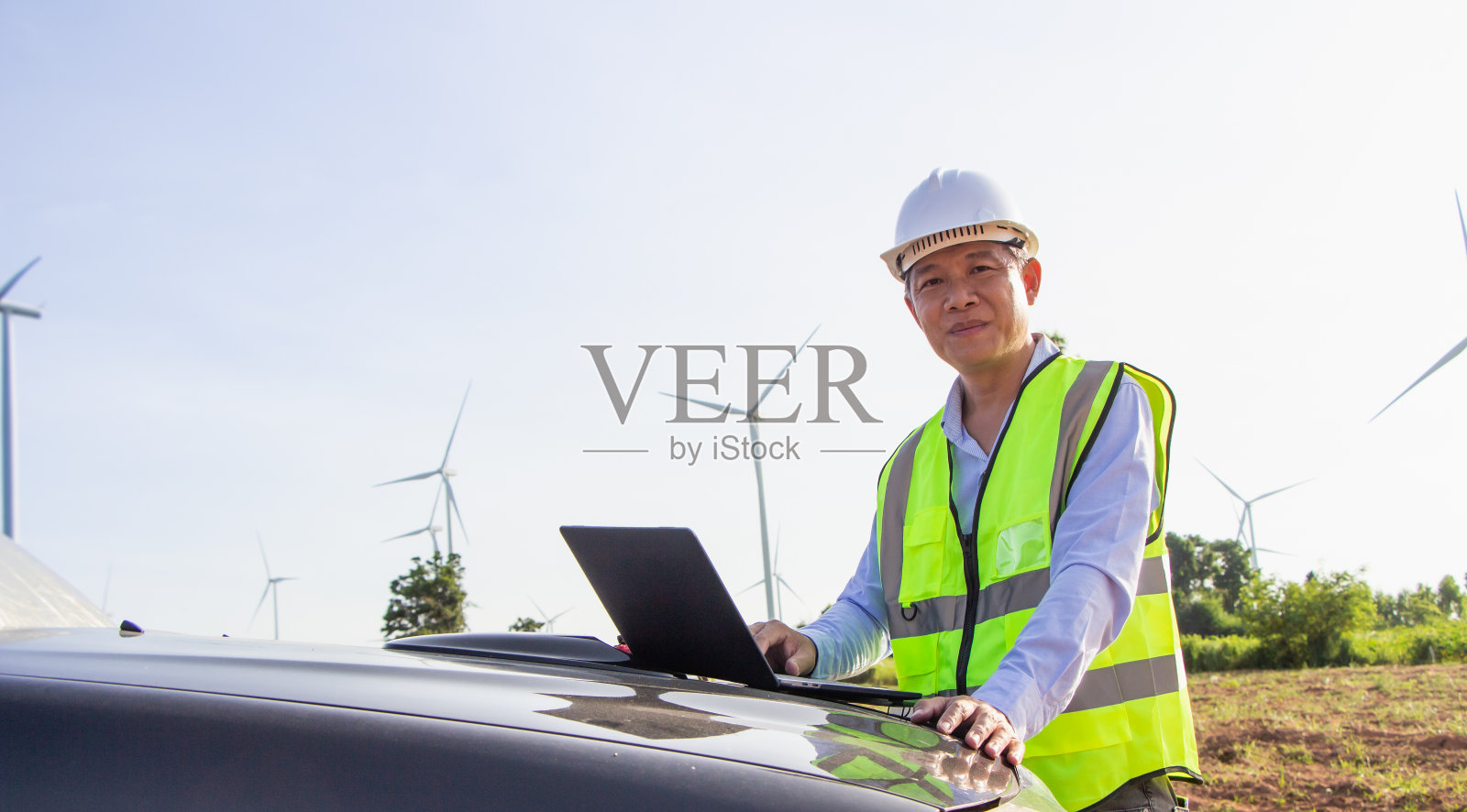 正面亚洲工程师微笑着拿着文件夹看着相机与风车，风力涡轮机的动力背景。照片摄影图片