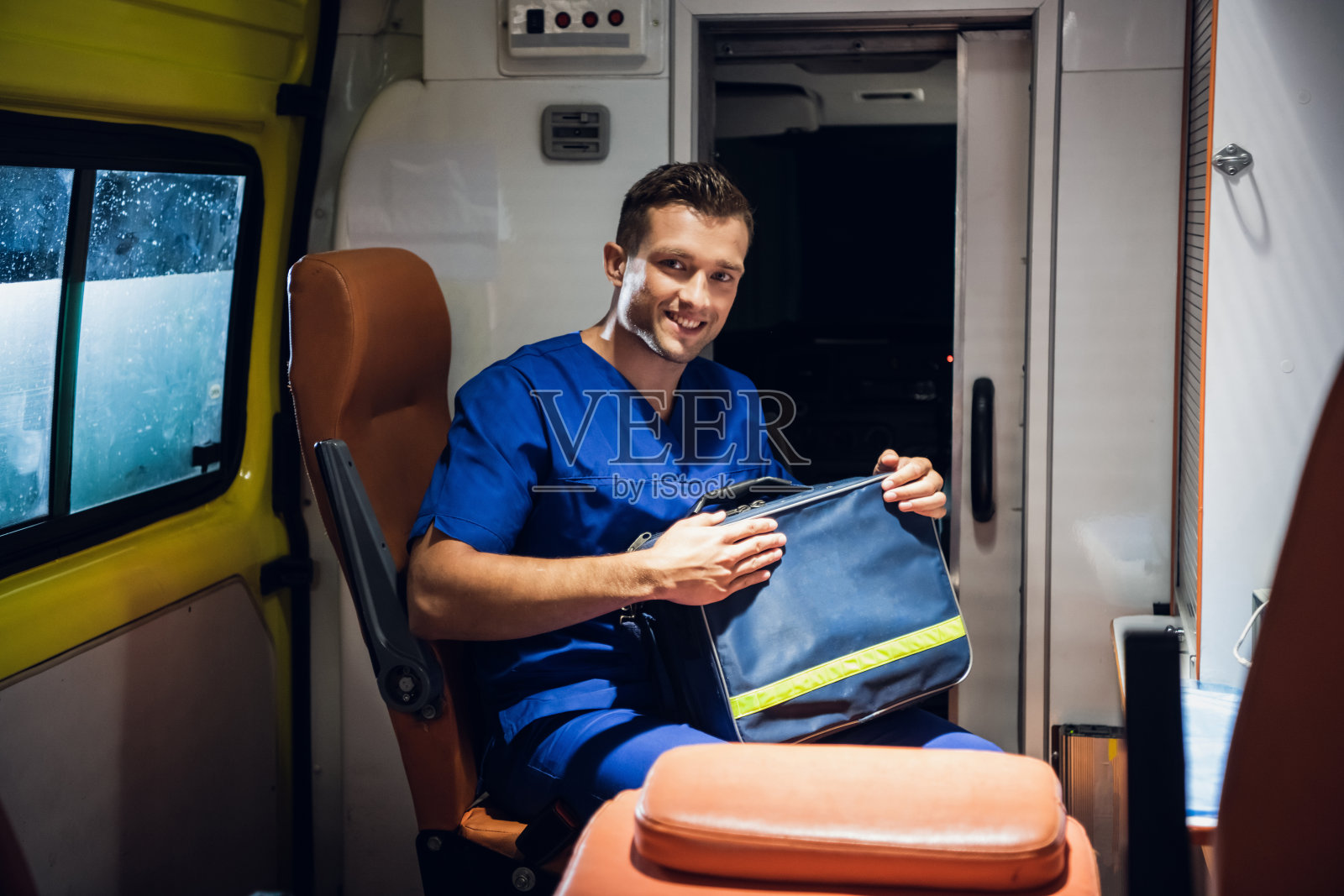 一位年轻的男性护理人员坐在救护车里，拿着他的医疗包，对着镜头微笑照片摄影图片