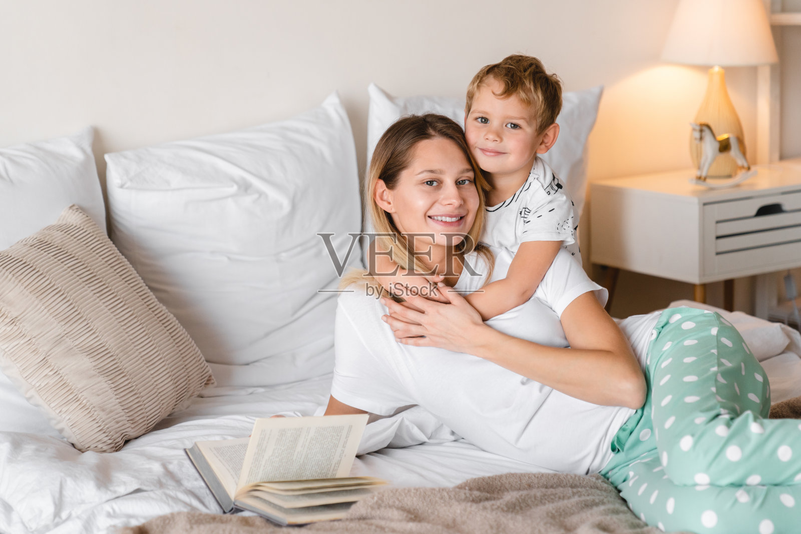 可爱的小男孩拥抱他年轻的妈妈在床上，而她正在给他读一本童话书。家庭时光，与孩子们共度时光照片摄影图片