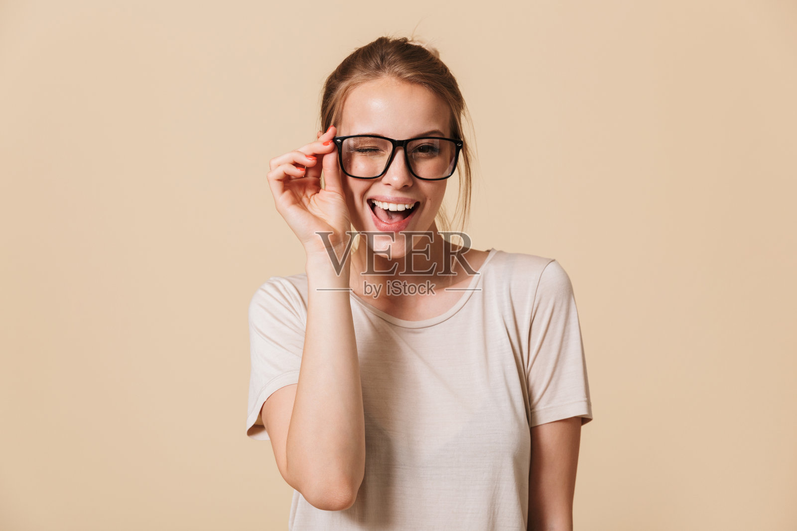 美丽开朗的女人20多岁的肖像，穿着基本的t恤，触摸眼镜，在镜头前微笑，同时眨眼，在工作室的米色背景孤立照片摄影图片