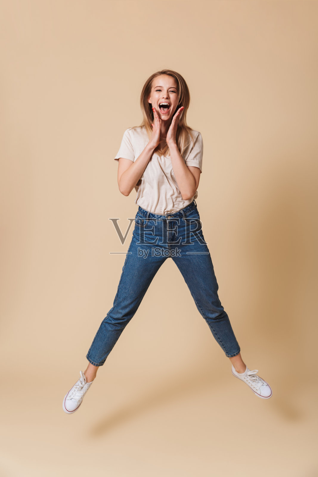 全身肖像快乐的金发女人20岁穿着休闲服装跳和鼓掌的手，孤立的米色背景照片摄影图片