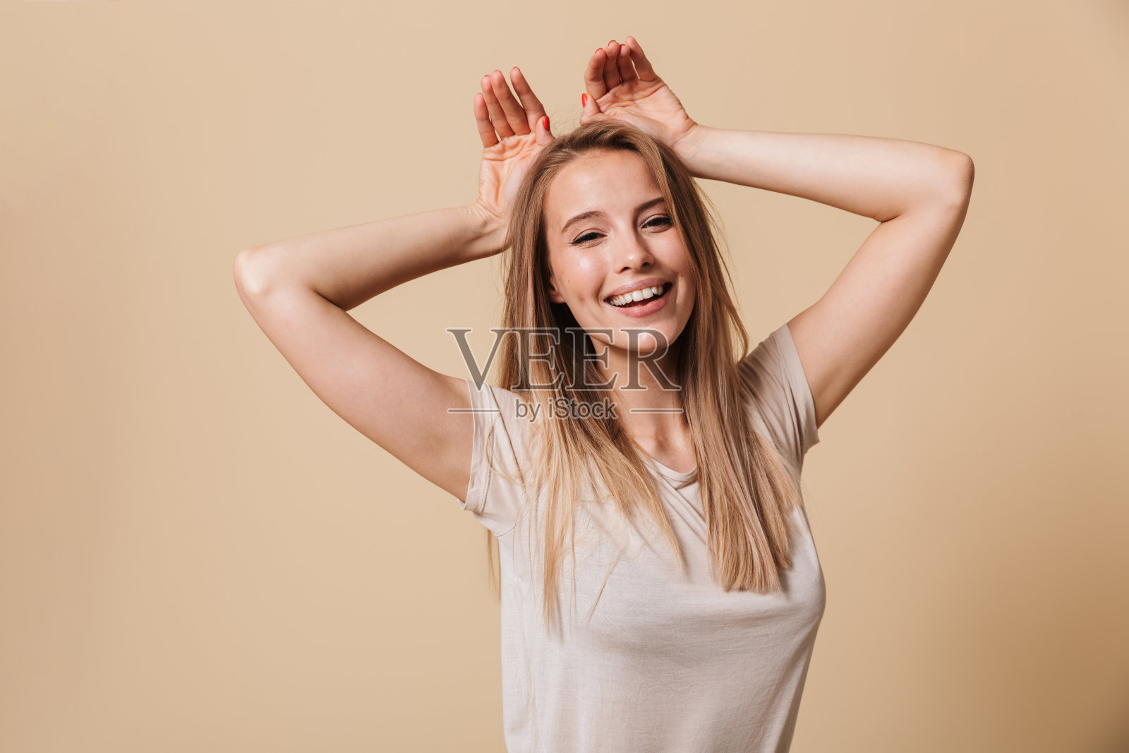 可爱的金发女孩20多岁的形象在休闲t恤微笑和显示兔耳朵在她的头，孤立在米色的背景照片摄影图片