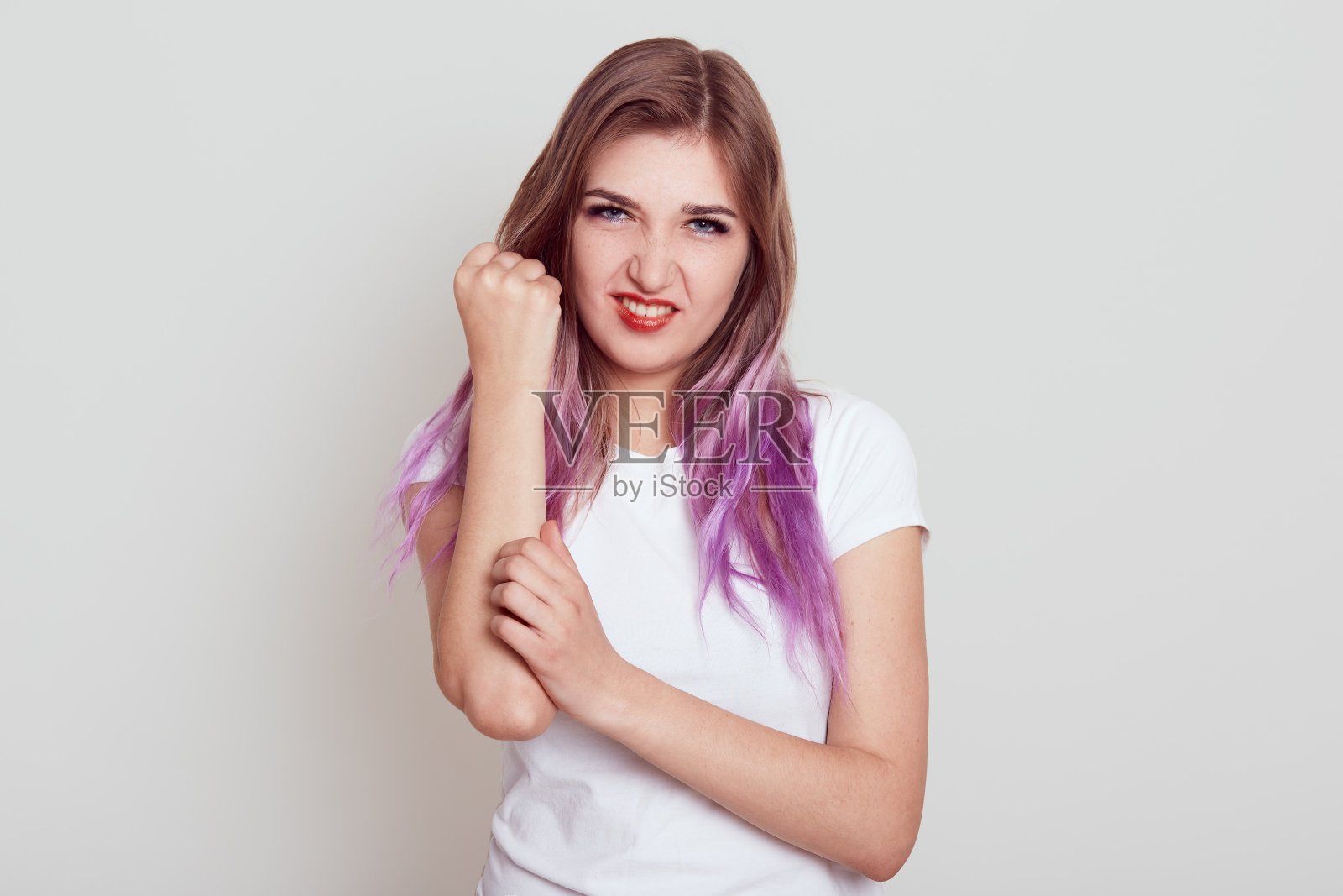 有魅力的年轻女孩淡紫色的头发伤害了她的手臂，触摸疼痛的手和直视镜头，有严重的伤害，孤立在灰色的背景。照片摄影图片