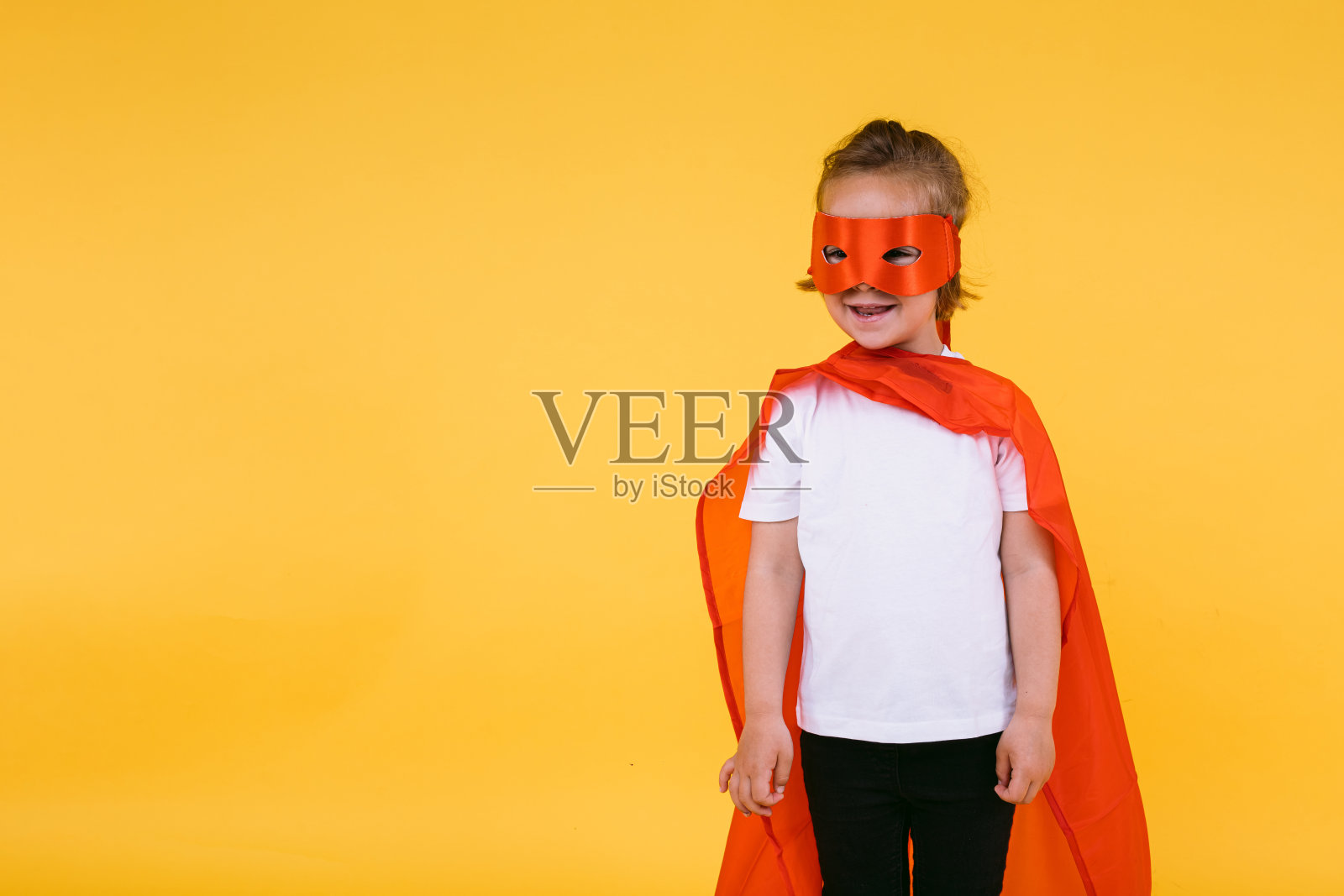 金发小女孩披着斗篷，戴着红色面具，装扮成超级英雄超级英雄，微笑着望向一边，在黄色的背景上照片摄影图片
