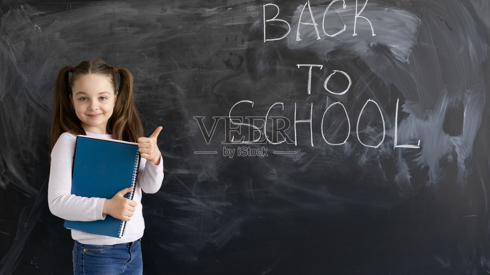 一个年轻的白人女学生站在黑板后面。这些字被写回学校。手里拿着几本大笔记本，大拇指朝上。照片摄影图片