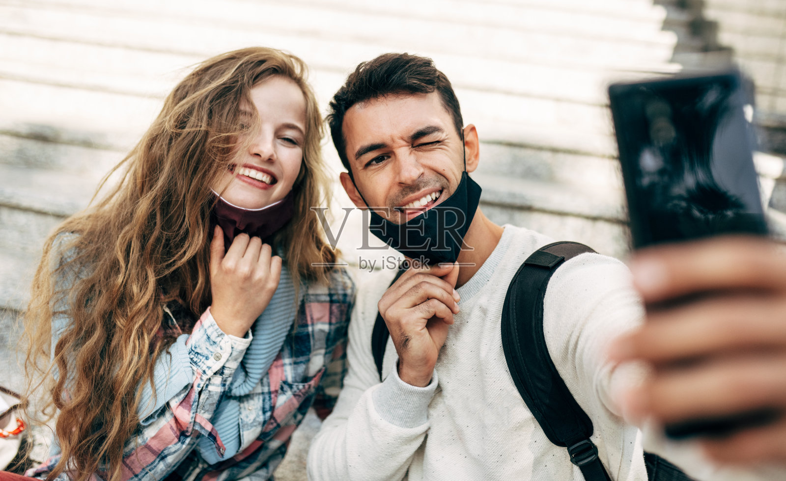 两个快乐的年轻学生坐在大学校园的楼梯上用手机自拍。带着防护面具的微笑女子和男子在智能手机上聊天。照片摄影图片