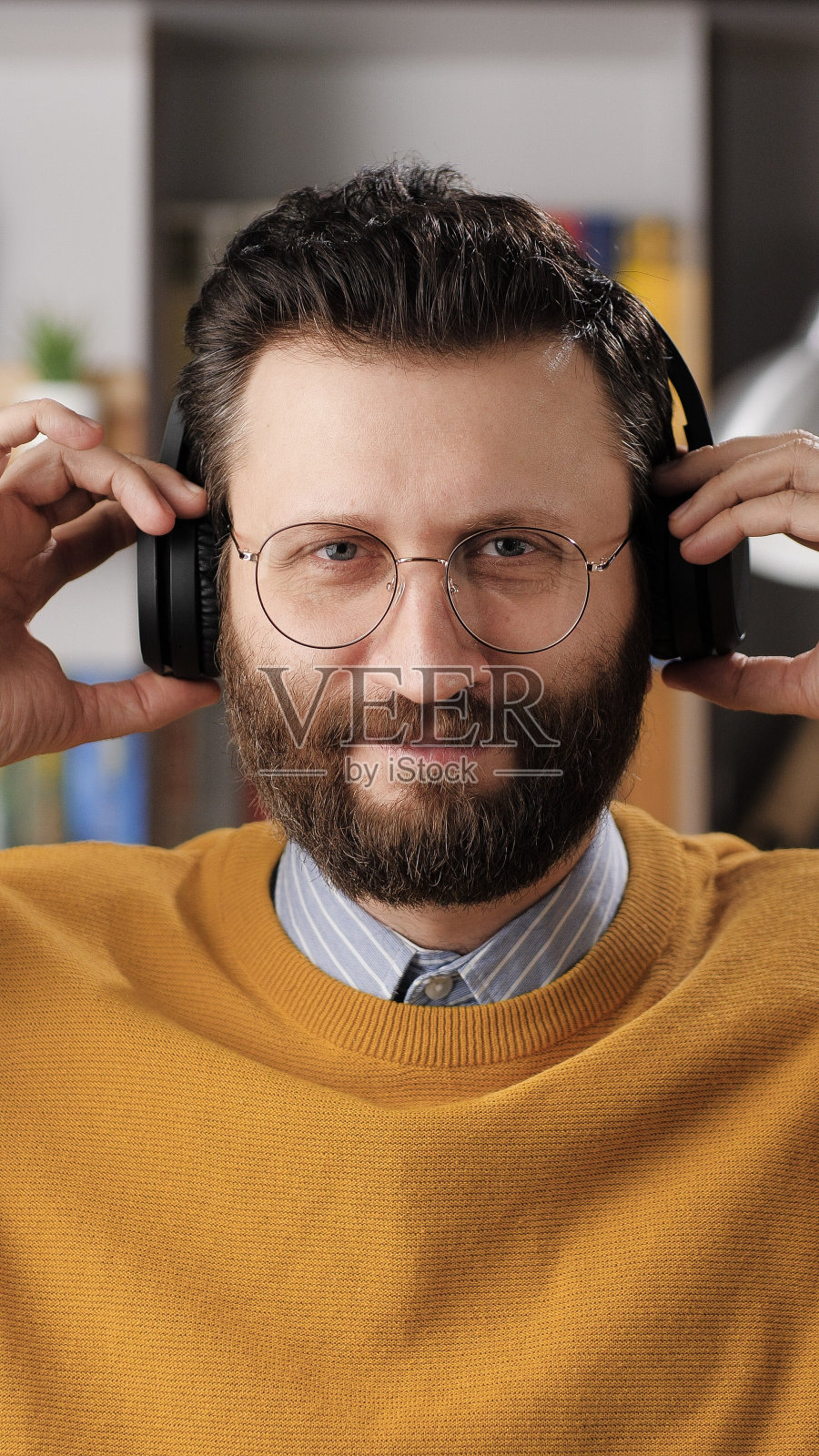 在线学习、远程教育、视频通话、e-learning、远程演示概念。垂直视角的正面大胡子男子看相机拿着无线耳机与他的手照片摄影图片
