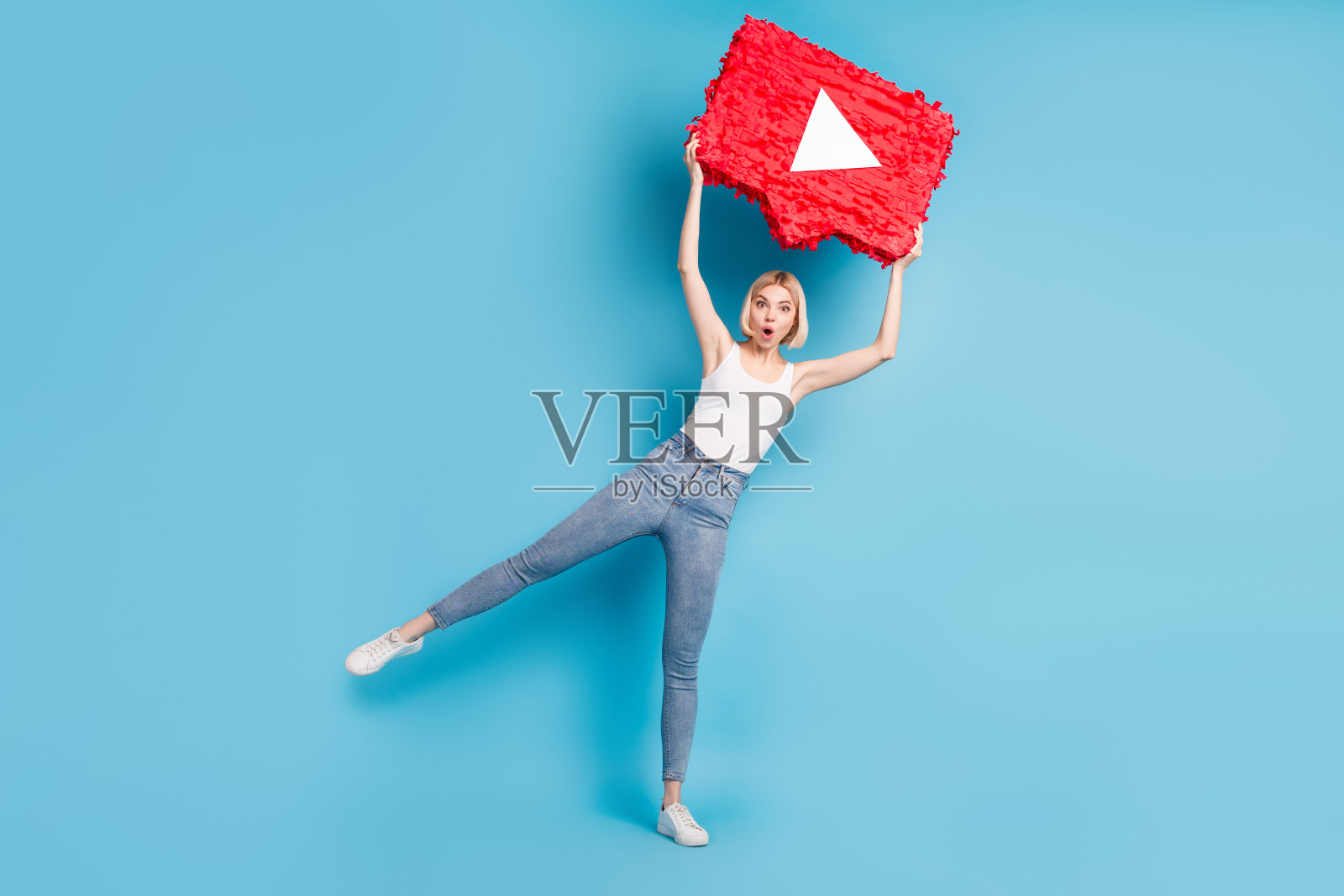 全长照片的惊奇震惊的年轻女子持有大的视频播放按钮，头顶上孤立的蓝色背景照片摄影图片