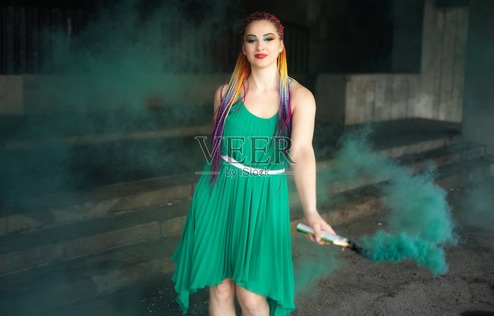 可爱的女孩在一个温暖的春天城市的街道上，穿着绿色的裙子，梳着非洲人的辫子，化着五颜六色的妆，与明亮的绿色烟雾摆姿势照片摄影图片