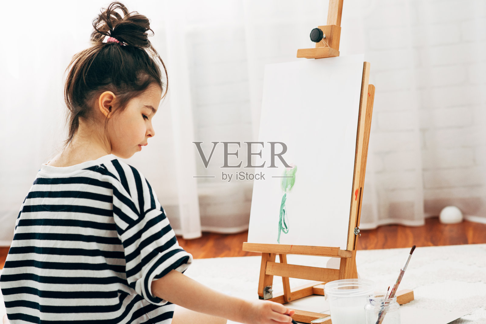 一个小女孩在家里的画架上画一朵花的横向后视图图像。可爱的孩子坐在地毯上，在她的房间里画画。照片摄影图片