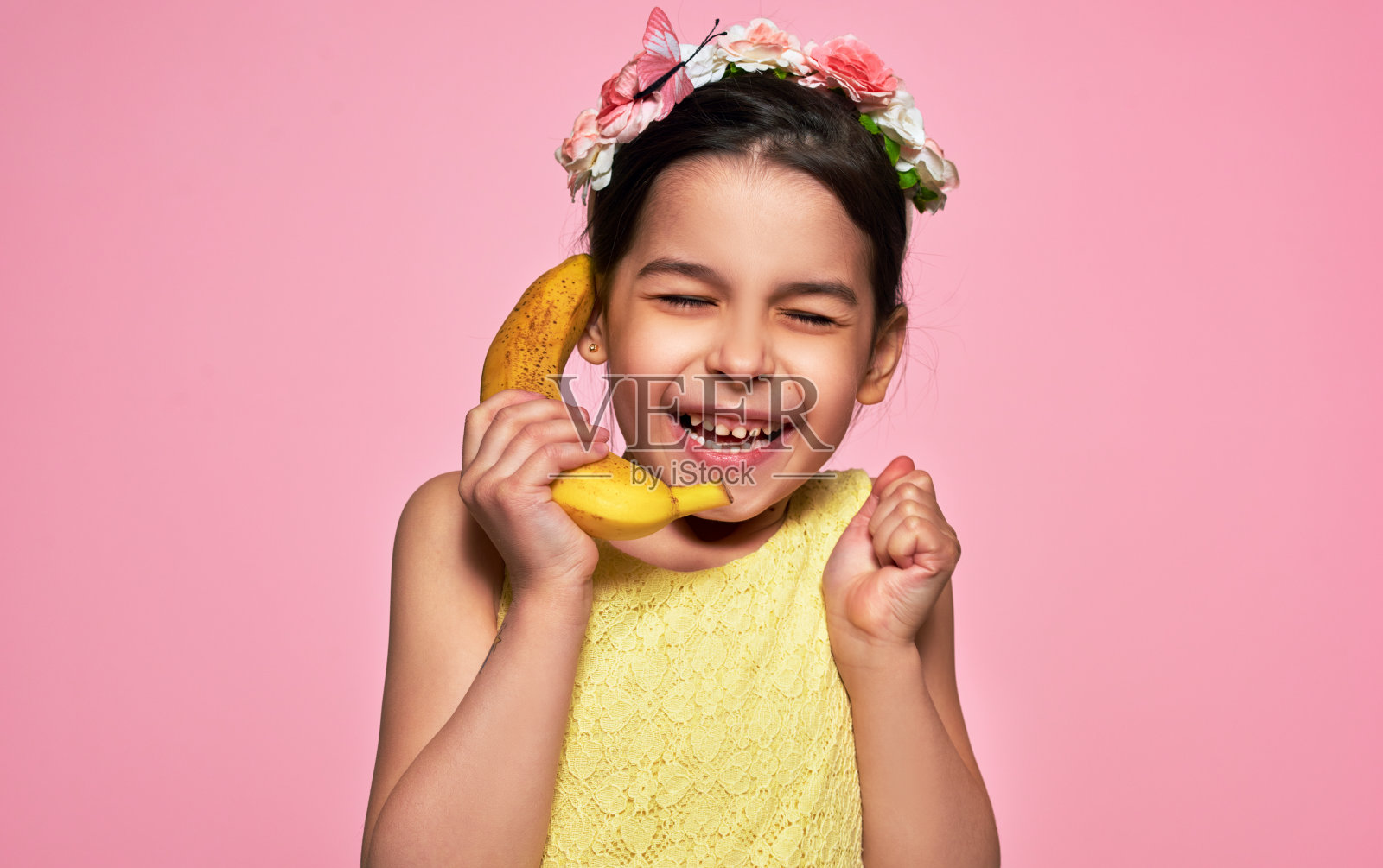 一个快乐的小女孩拿着香蕉，像手机一样，孤立在粉红色的背景下。一个穿着黄色连衣裙的快乐的孩子假装在对着香蕉说话，就像在打手机。照片摄影图片