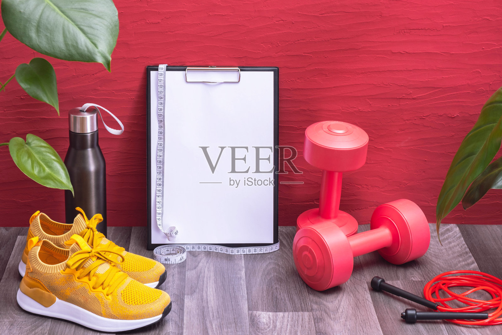 在家锻炼-红色哑铃，水瓶和跳绳，笔记板和卷尺，黄色运动鞋和大绿叶，红色背景照片摄影图片