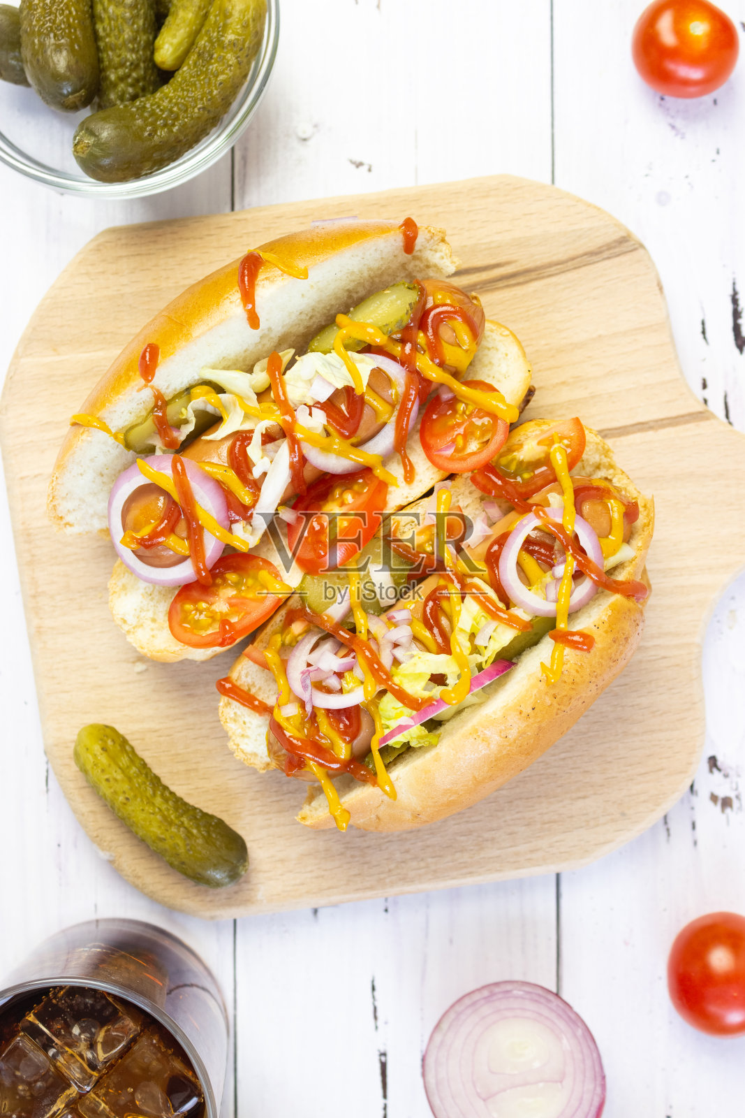 白色木桌上的切菜板上放着热狗、蔬菜、芥末和番茄酱，俯视图。蔬菜和饮料。快餐，街头小吃照片摄影图片