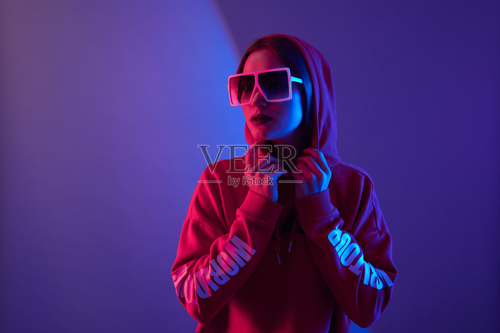 时尚年轻女子模特穿帽衫和眼镜太阳镜在霓虹灯工作室的背景。照片摄影图片