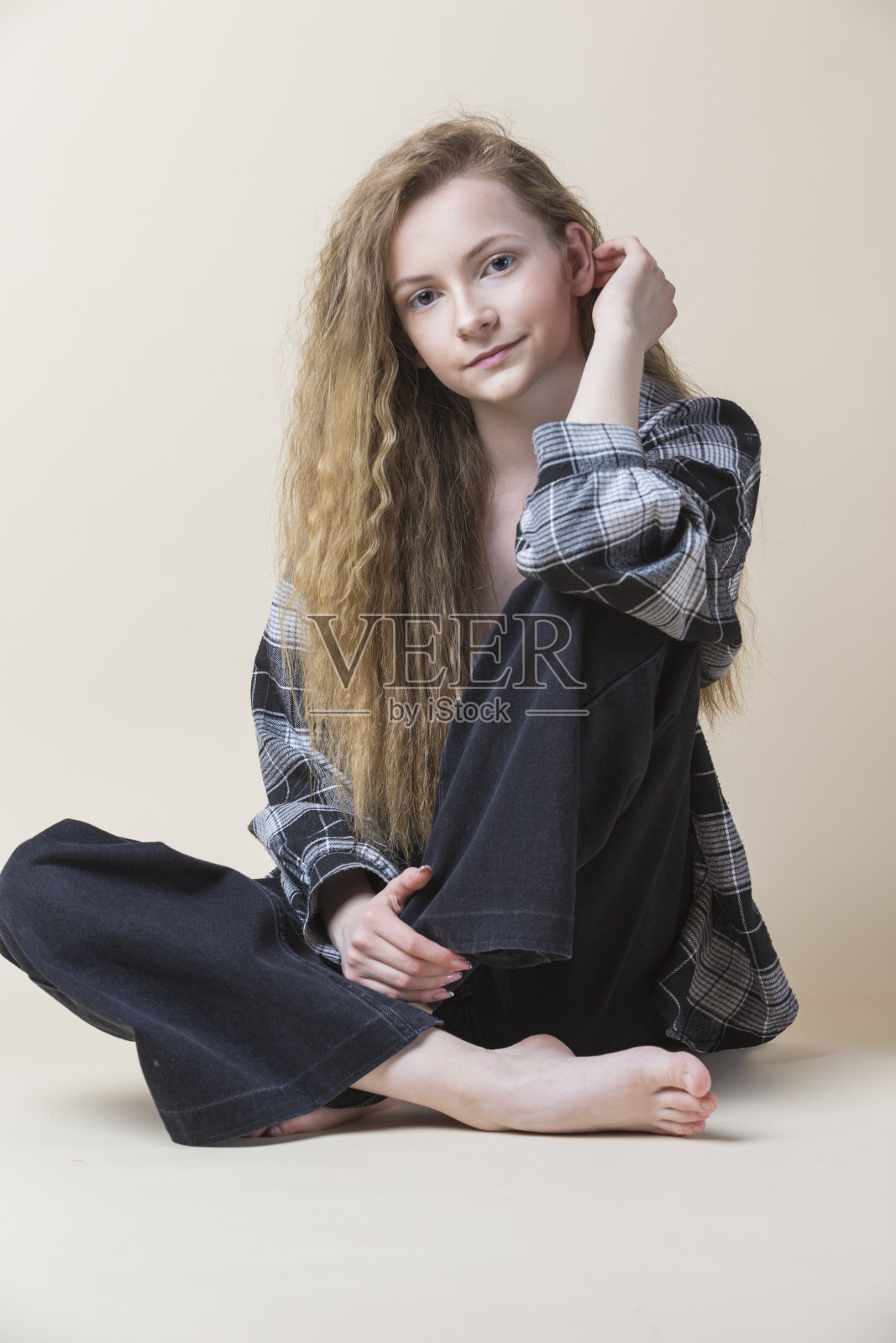 穿着黑白格纹衬衫，盘腿坐在米色背景前的白人金发少女。竖版构图照片摄影图片