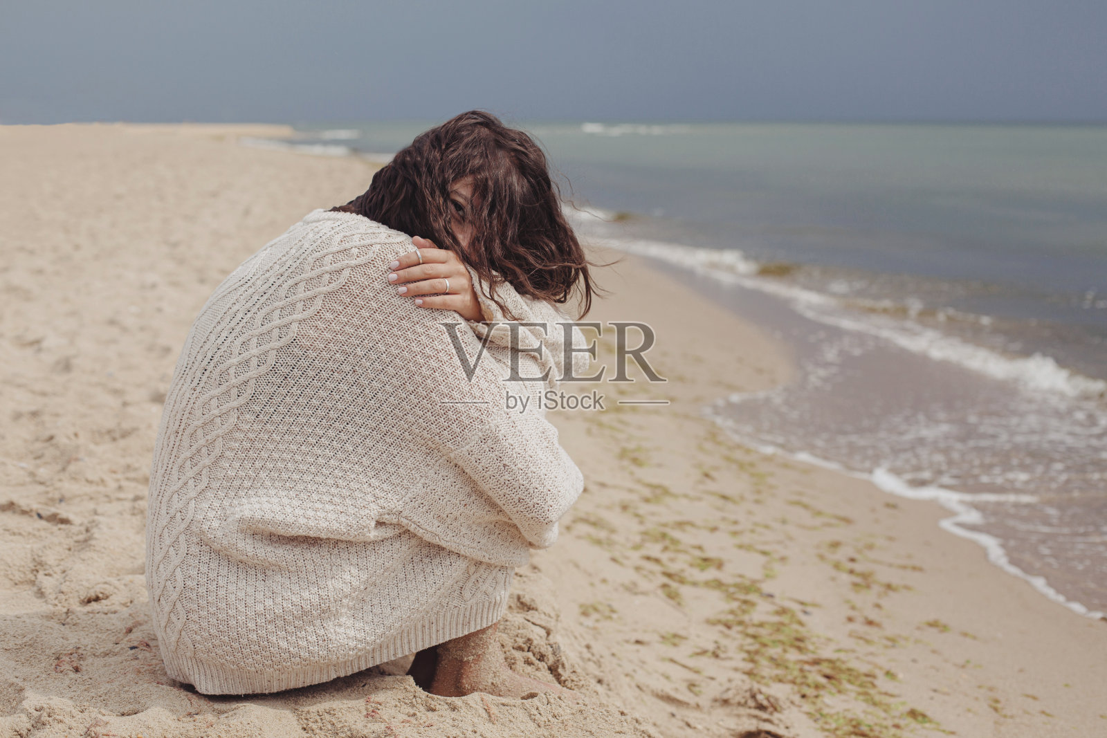 无忧无虑的美丽女子带着随风卷曲的头发坐在海边的沙滩上，平静的时刻。时尚年轻快乐的女性在针织毛衣放松海岸。暑假的心情照片摄影图片