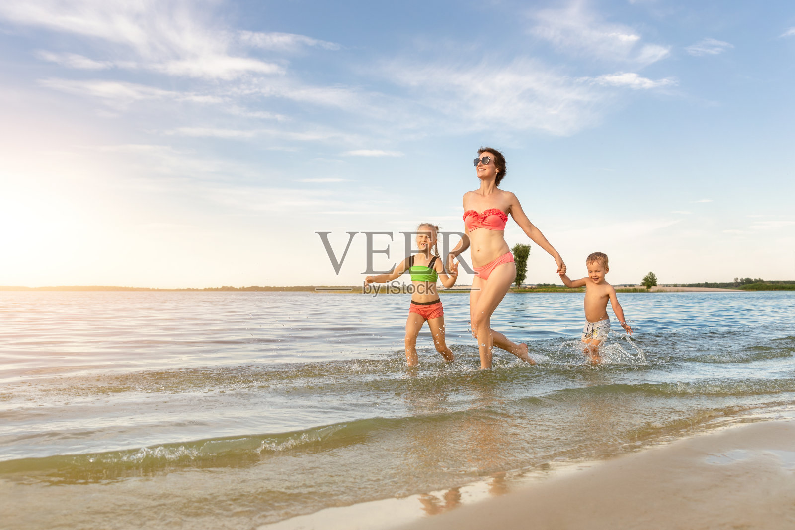 年轻的成年人有吸引力的苗条运动母亲享受乐趣，在湖或海沙缺口与两个可爱的小兄弟姐妹对蓝色的天空在夏天的一天。夏季家庭度假概念照片摄影图片