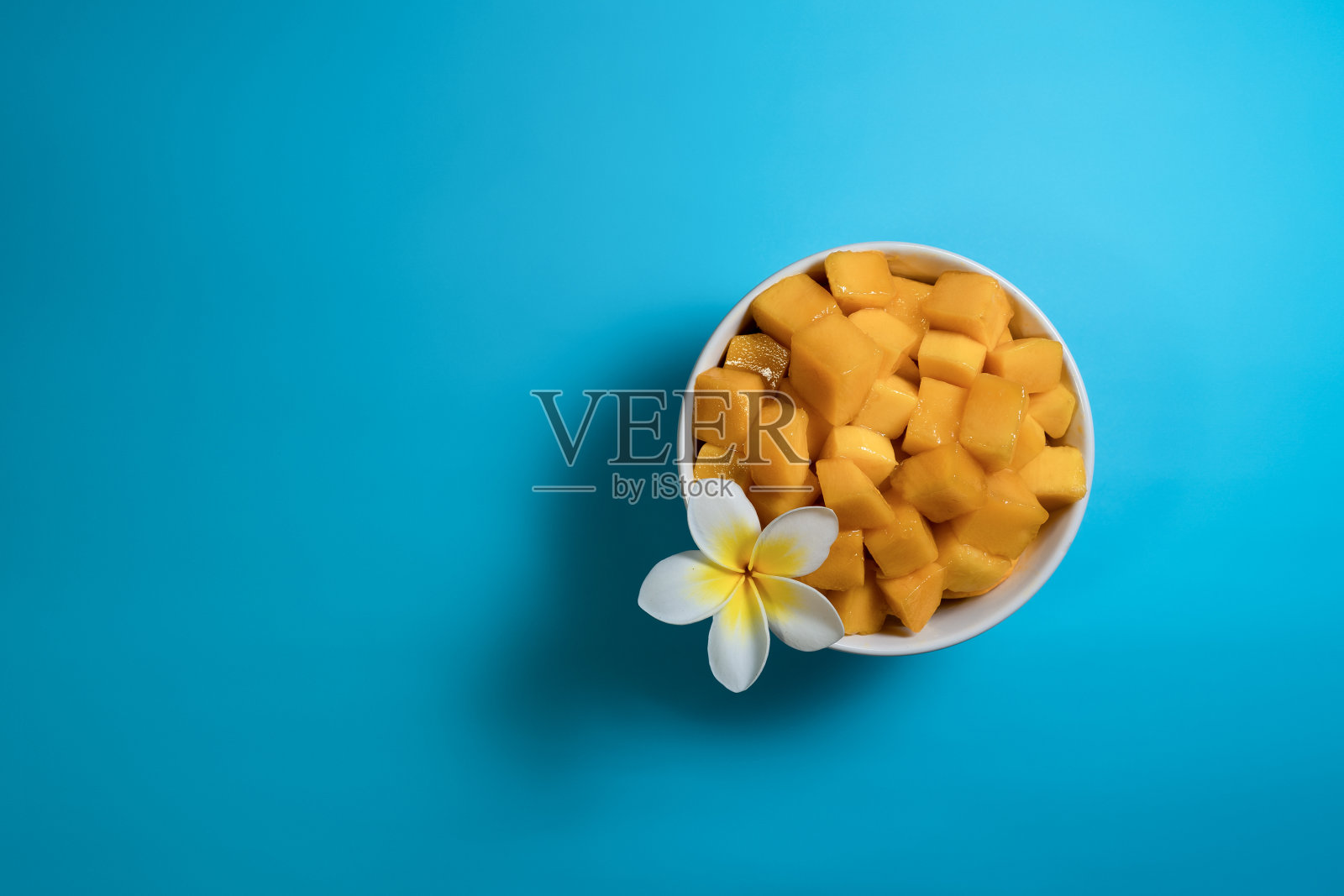 热带水果的概念。白色的碗里放着黄色的芒果块，背景是亮蓝色的，花是白色的。平铺，俯视图，拷贝空间照片摄影图片