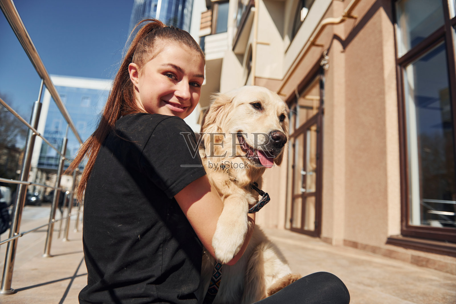 坐在一起。年轻积极的女人有乐趣与她的狗当有一个户外附近的商业大楼照片摄影图片
