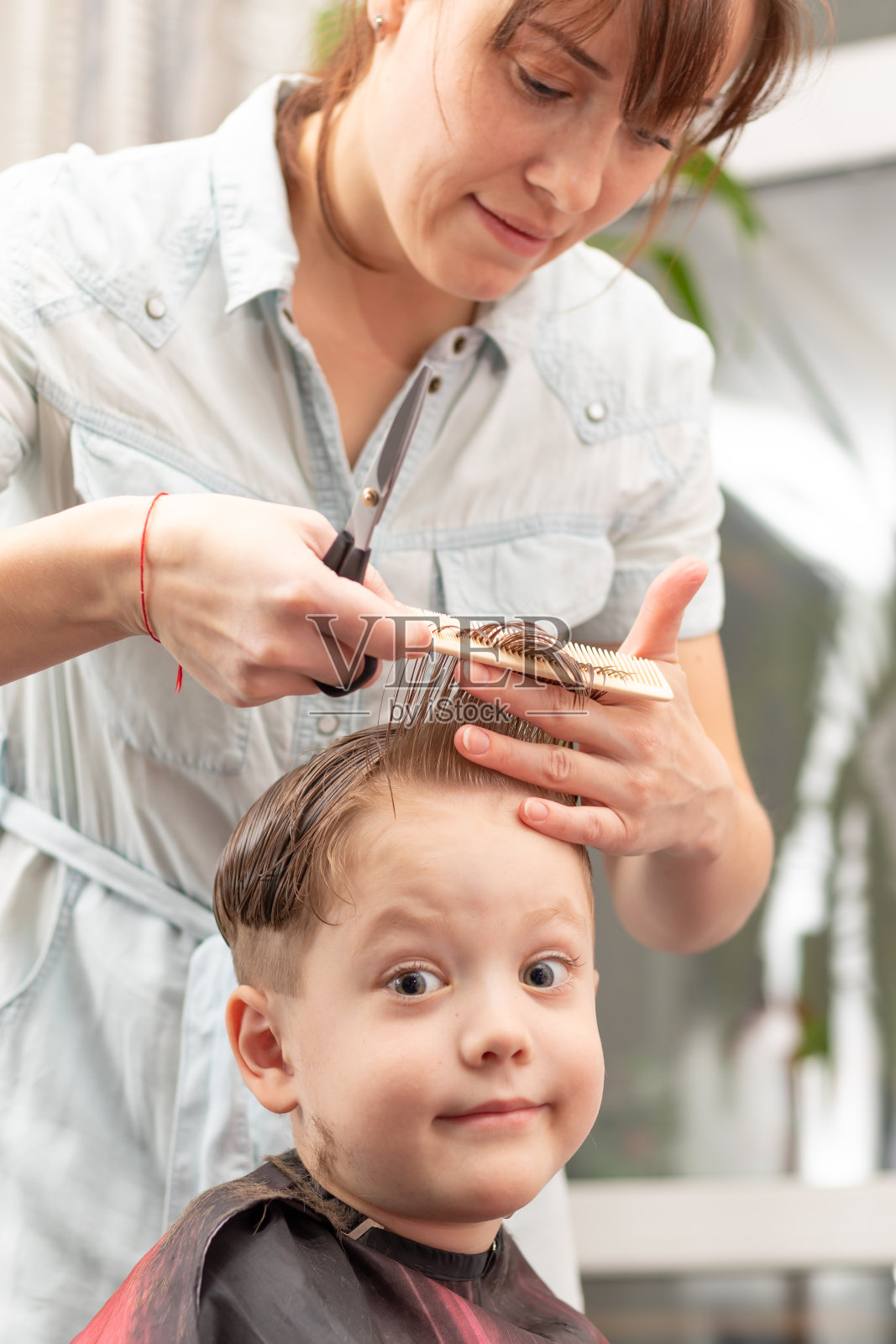 年轻漂亮的妈妈穿着浅蓝色的衣服，手里拿着梳子和剪刀在家给儿子理发。有选择性的重点照片摄影图片