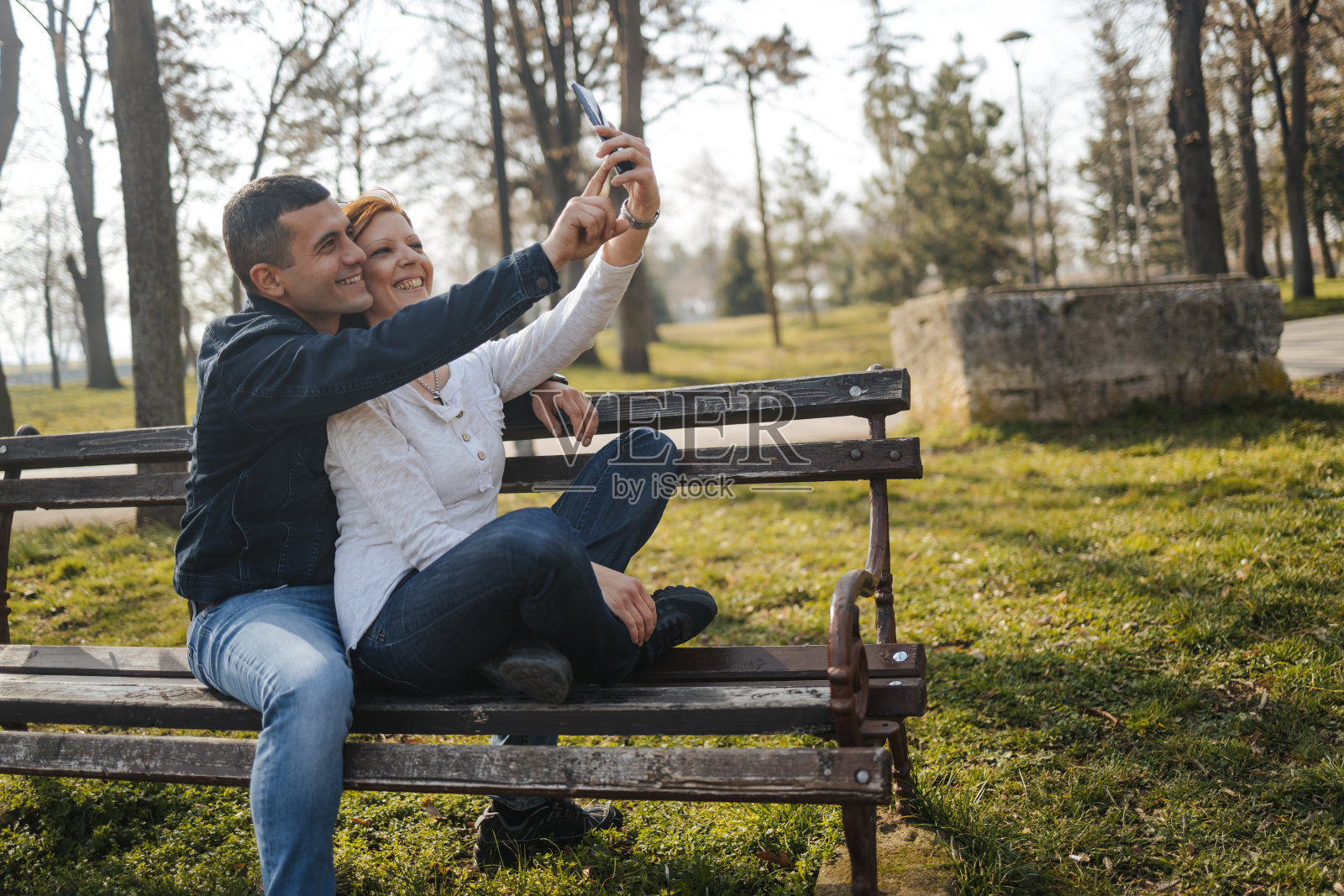 一对幸福的年轻情侣坐在公园的长椅上，用手机自拍照片摄影图片