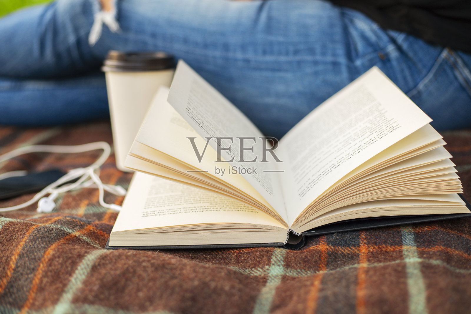 在公园里，一个穿着牛仔裤、拿着一本书、一杯咖啡和一部带耳机的智能手机的女人坐在毯子上。照片摄影图片