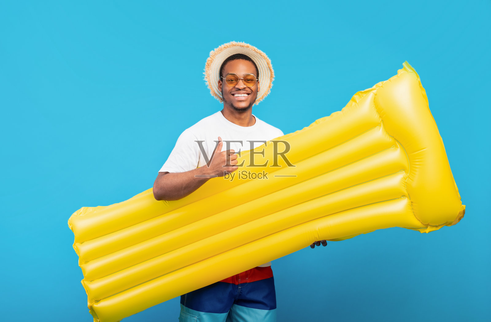 年轻的黑人小伙子夏天戴着草帽，拿着黄色的充气气垫，在蓝色的背景上竖起大拇指照片摄影图片