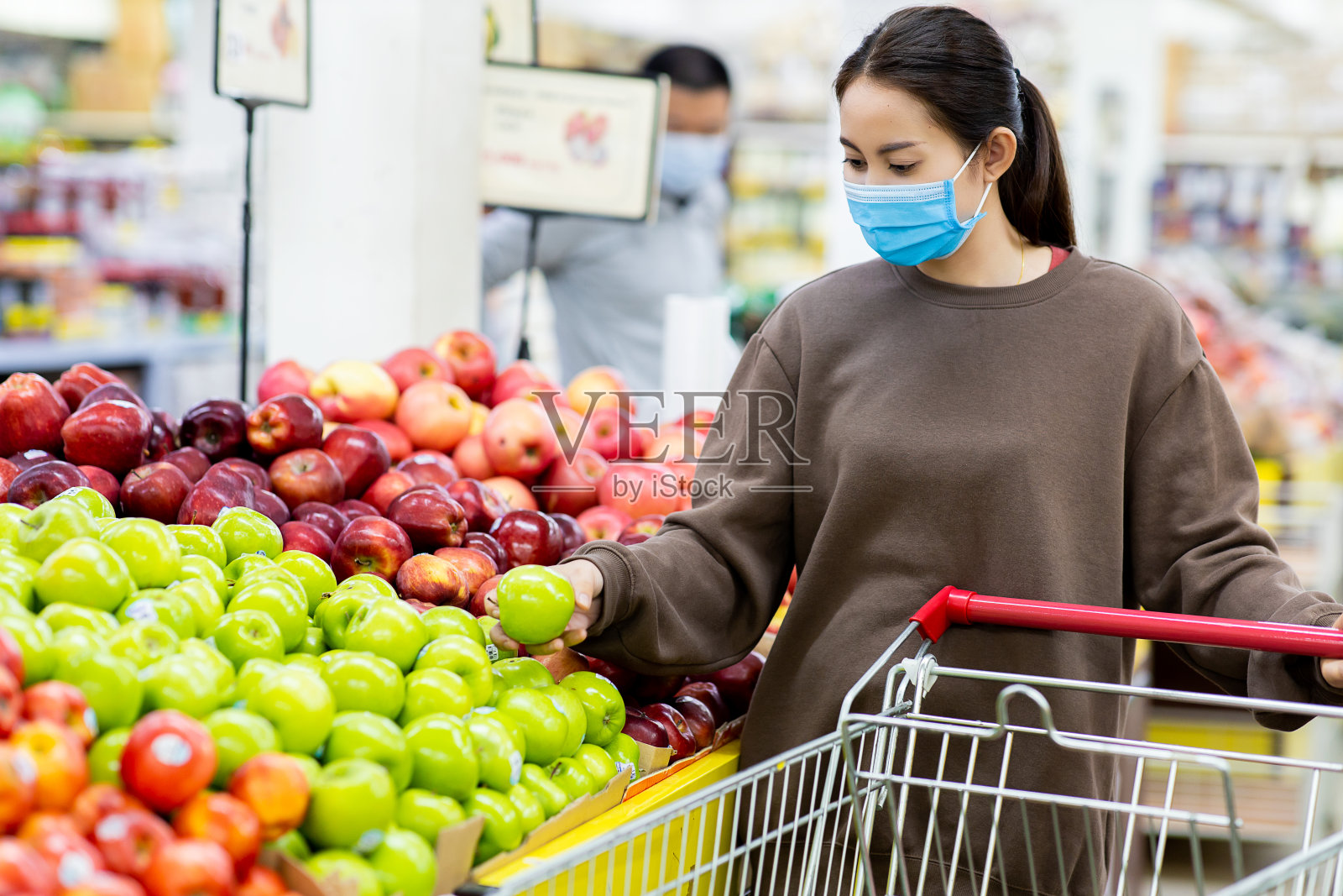 新冠肺炎疫情期间，年轻的亚洲女性戴着防护口罩在超市推着购物车购买新鲜水果。照片摄影图片