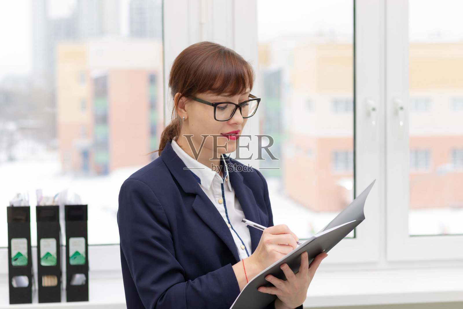 年轻漂亮的女商人戴着眼镜，在窗边一间光线明亮的办公室里，手里拿着一个文件夹在做笔记照片摄影图片