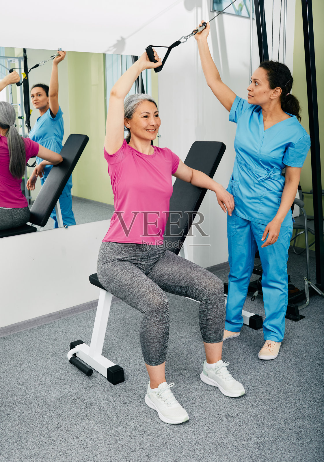 一名中国妇女坐在运动长椅上，在医学运动中心她的康复医生附近使用拉绳健身器进行康复照片摄影图片