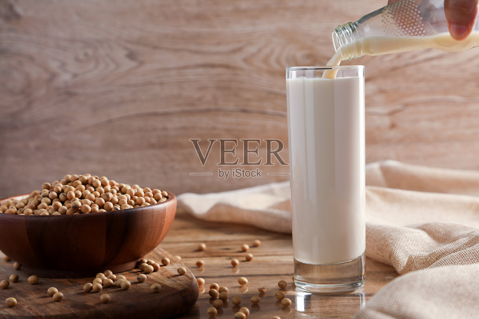 将豆浆倒入玻璃杯中，以大豆为背景倒在木桌上。健康食品和素食的概念。照片摄影图片