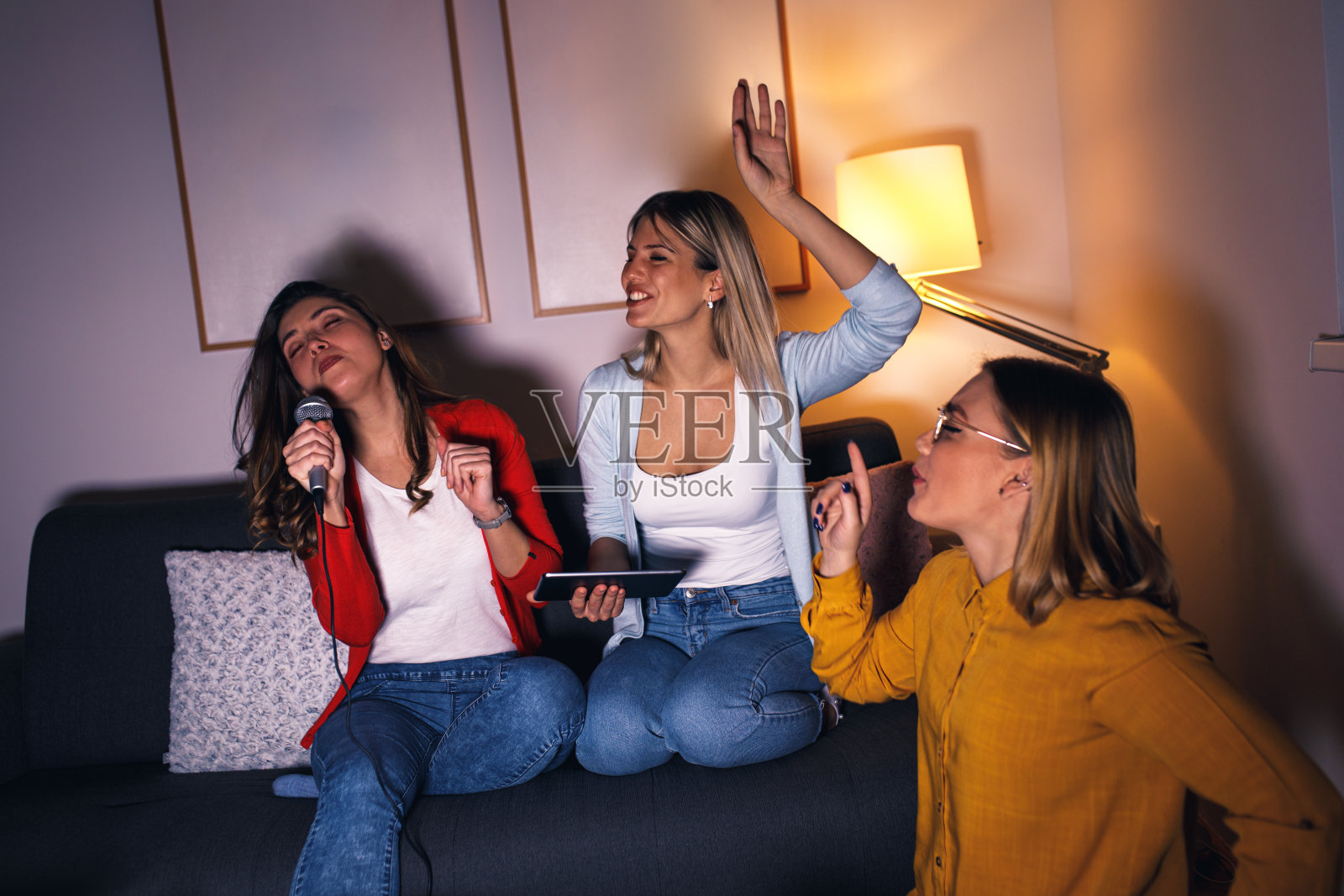 三个女性朋友坐在沙发上玩卡拉ok在家。照片摄影图片
