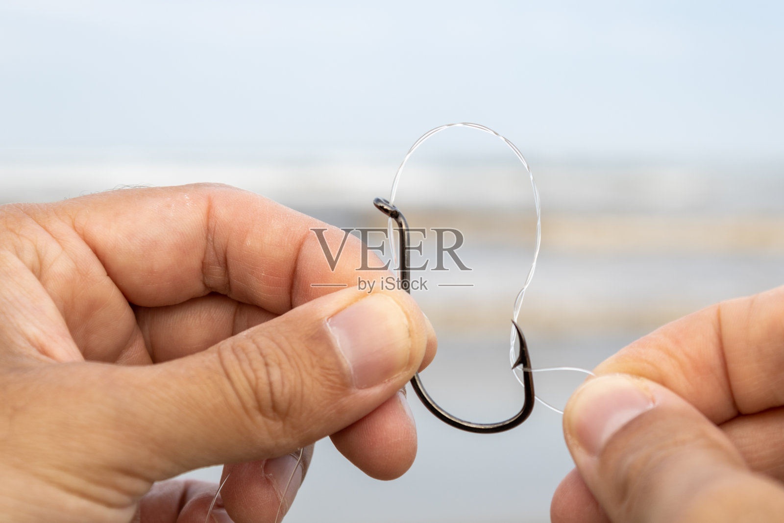一个白人的手在大海前用尼龙钓鱼线把一个黑色鱼钩的眼睛钓进去。提到在阳光明媚的周末与家人一起钓鱼、运动和休闲。照片摄影图片