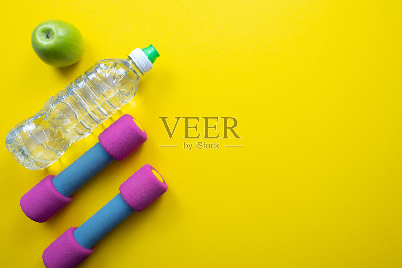 黄色背景——粉色哑铃，一瓶水和一个绿苹果，背景的右边是一个特写照片摄影图片