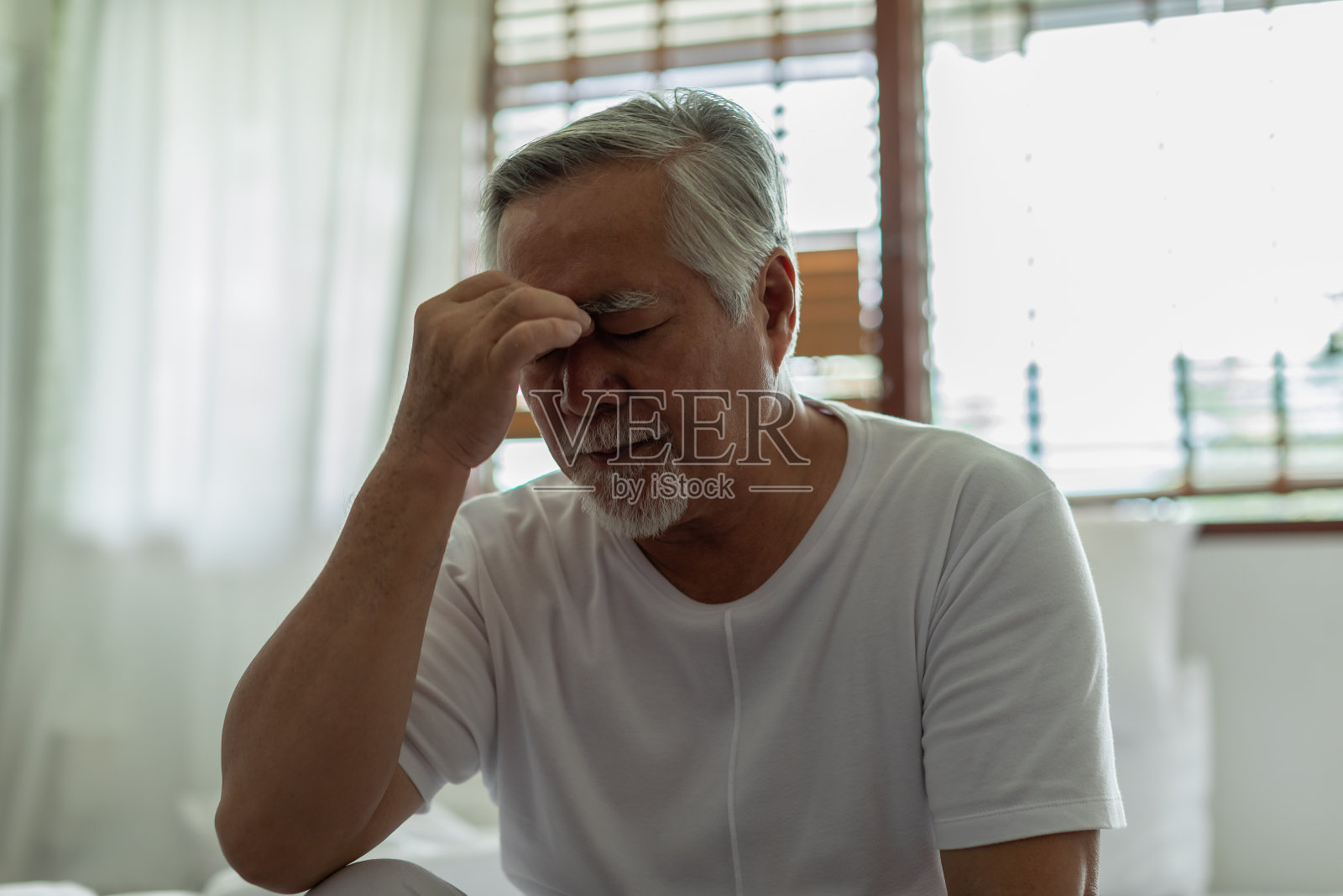 亚洲老年人白发头痛和偏头痛所以疼痛，压力老年人独自坐在家里的床上，不开心的老年人碰着头严重和尝试，不开心的老年人沮丧的概念，低调照片摄影图片