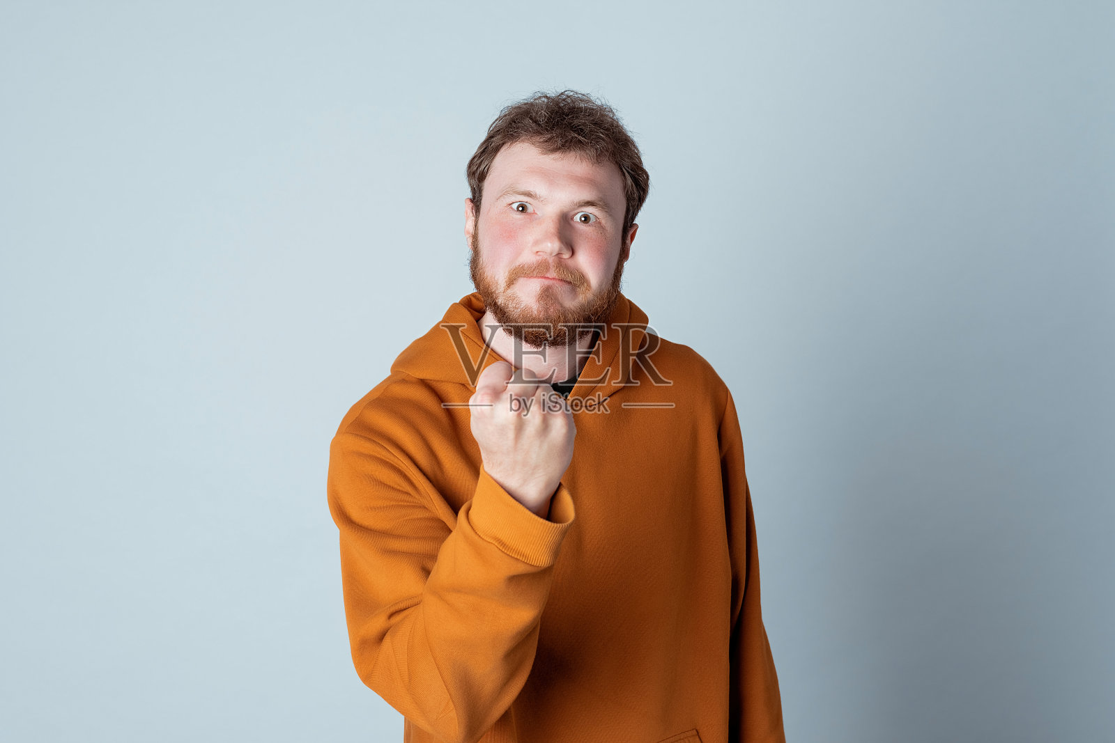 一名愤怒的蓄着胡须的年轻人，穿着时髦的连帽衫站在片场灰色的背景下，一边握拳，一边看着镜头照片摄影图片