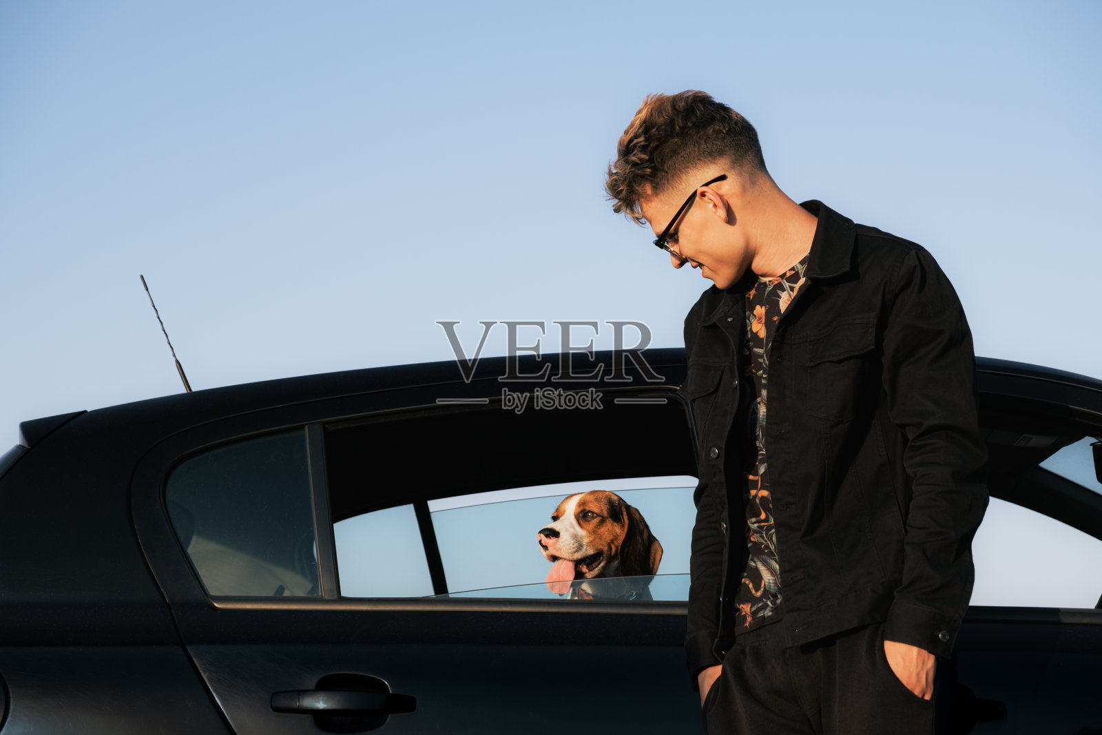 一个年轻人站在车旁，一只小猎犬从车窗往外看。照片摄影图片