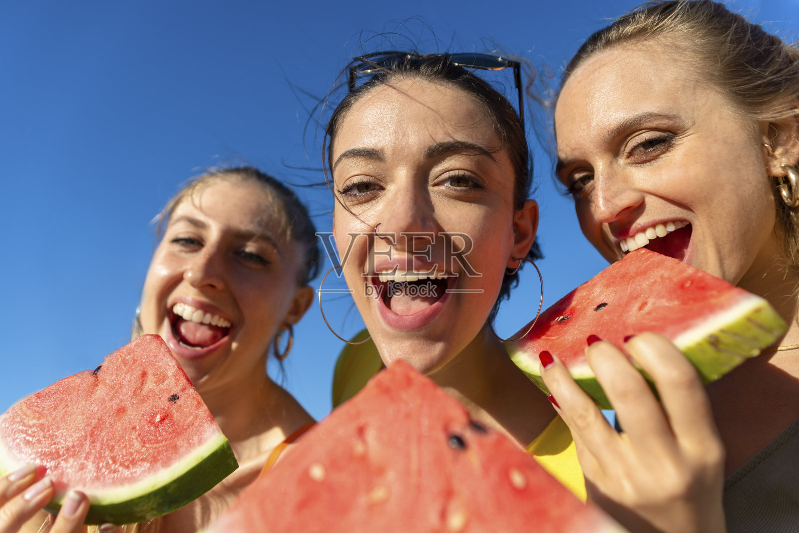 在夏天的时候，三个快乐的女孩在户外的海滩上吃西瓜。朋友们有有趣的孤立背景享受和咬新鲜的水果在海边度假。贞照片摄影图片
