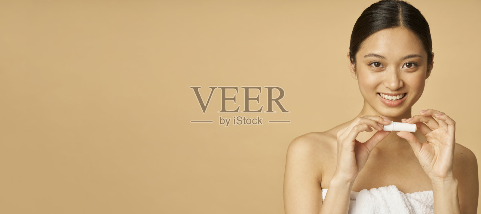 照片的网站标题，可爱的年轻女子包裹在毛巾微笑着，同时拿着唇膏，在米色的背景下摆出孤立的姿势照片摄影图片