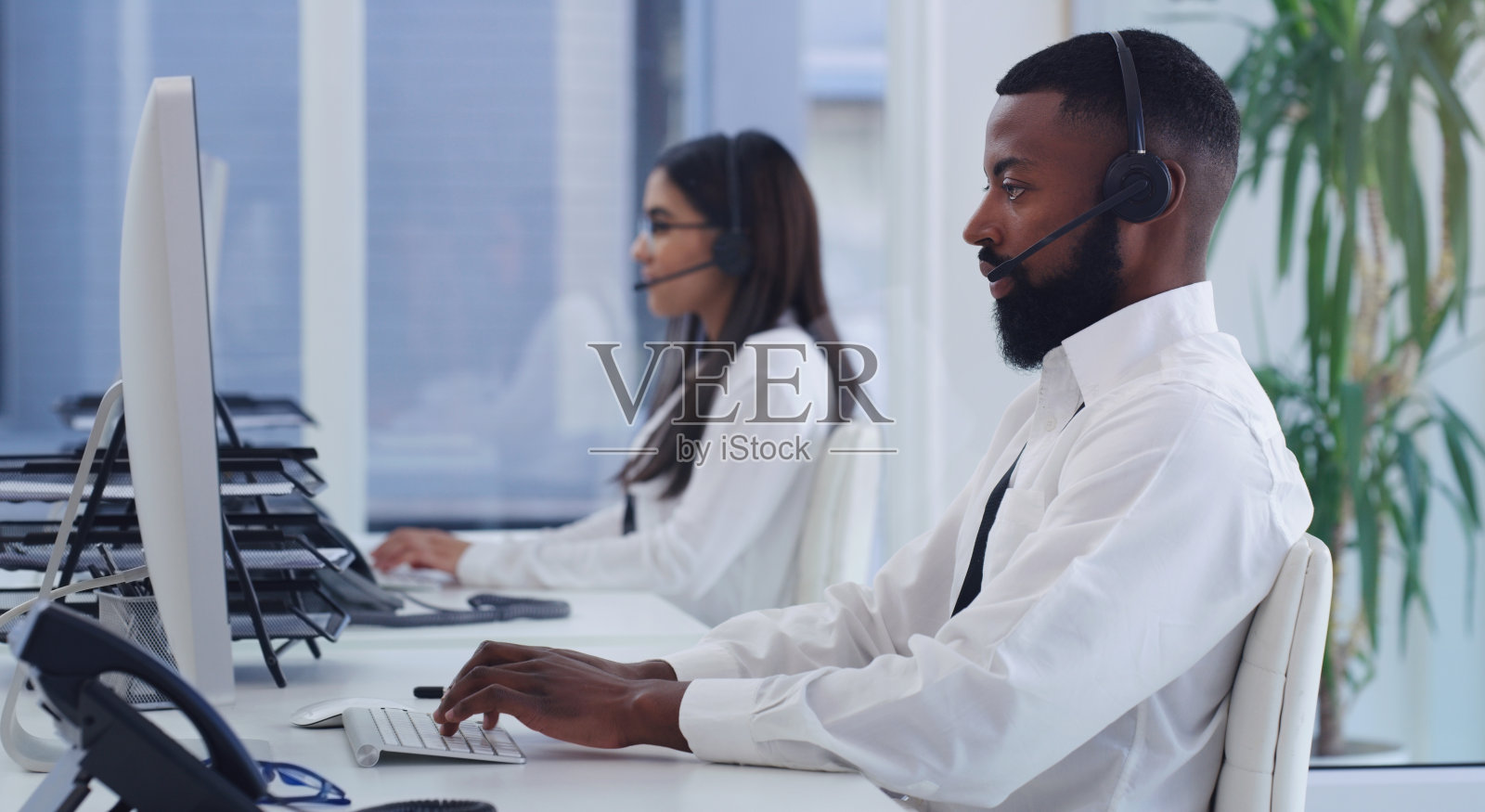 这是一个年轻的商人在呼叫中心用电脑工作的照片，背景是一位同事照片摄影图片