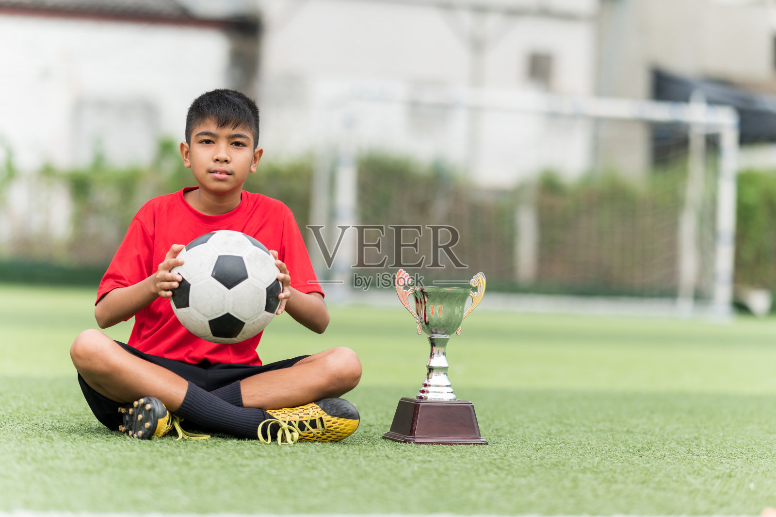 亚洲少年足球运动员坐在足球场上，旁边放着足球和奖杯。照片摄影图片