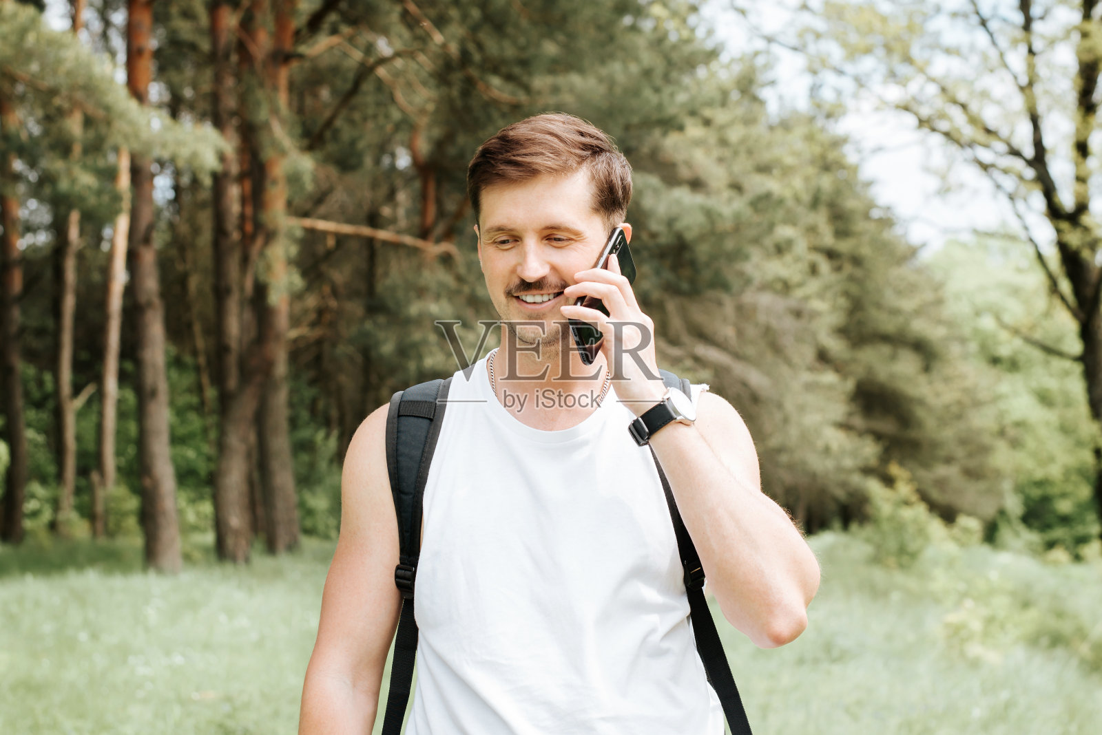 微笑的年轻人在树林里走路时用手机聊天的肖像。快乐的男人背着双肩包在大自然中远足，愉快的呼唤户外。照片摄影图片