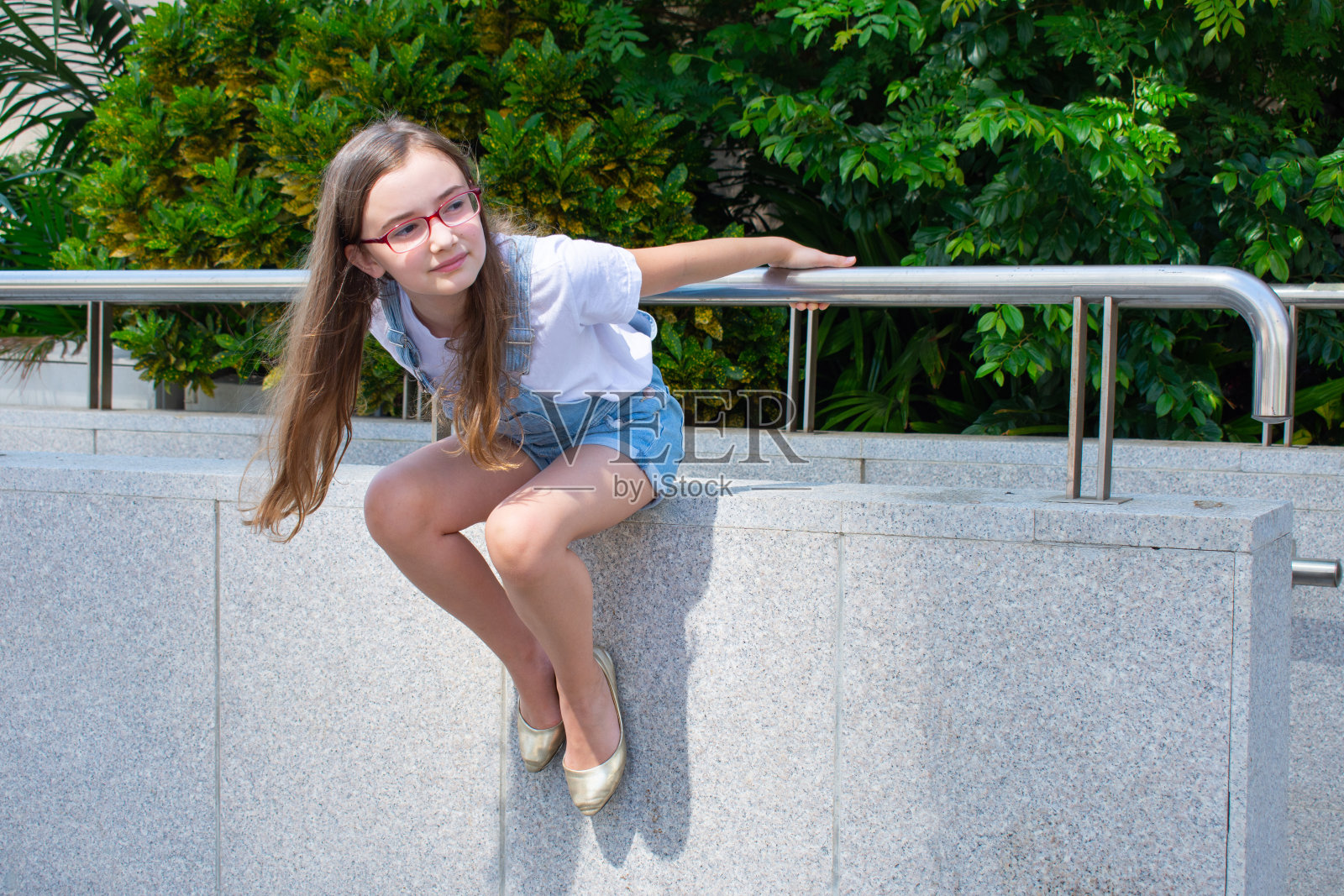 戴眼镜、穿着白色T恤和牛仔背带裤的女孩在阳光明媚的天气里坐在栏杆上照片摄影图片