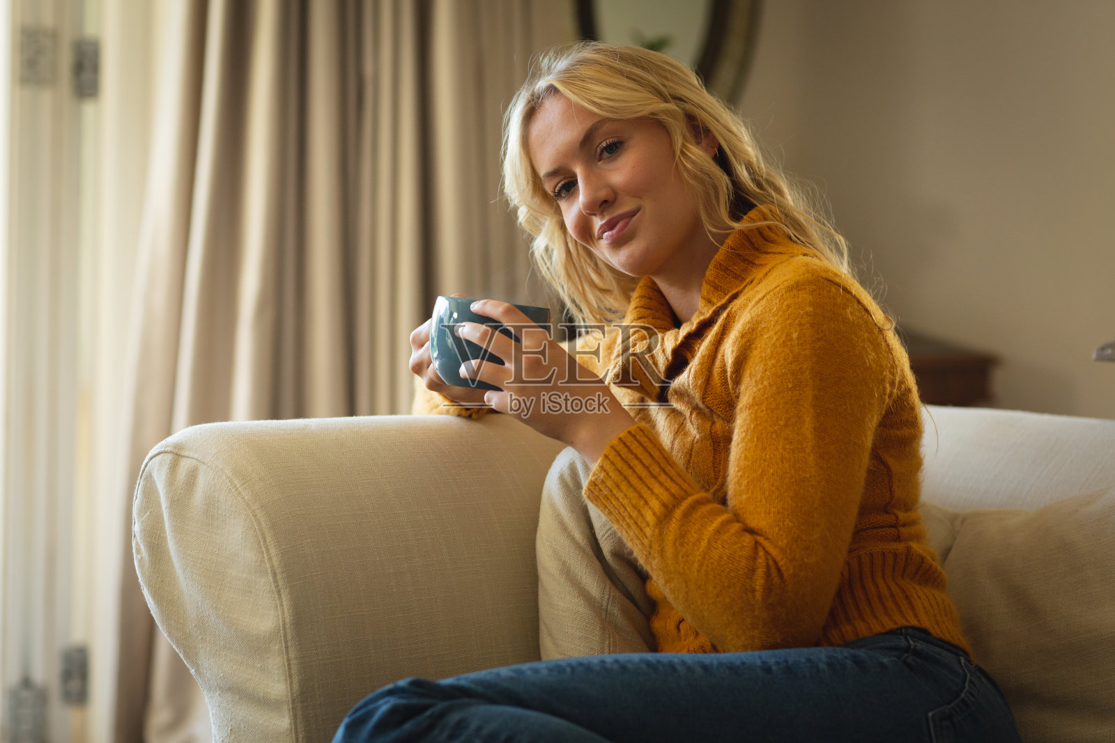 一个快乐的白人女人坐在客厅的沙发上，拿着咖啡杯，微笑着照片摄影图片