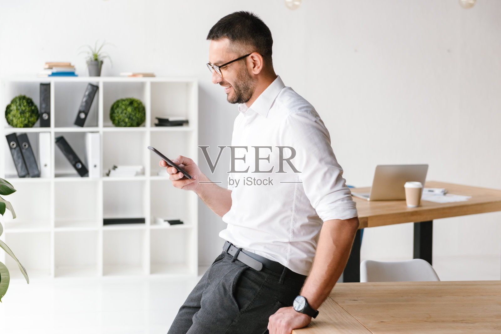 英俊快乐的男人穿着白衬衫坐在办公室的桌子上，并使用智能手机进行商务聊天照片摄影图片