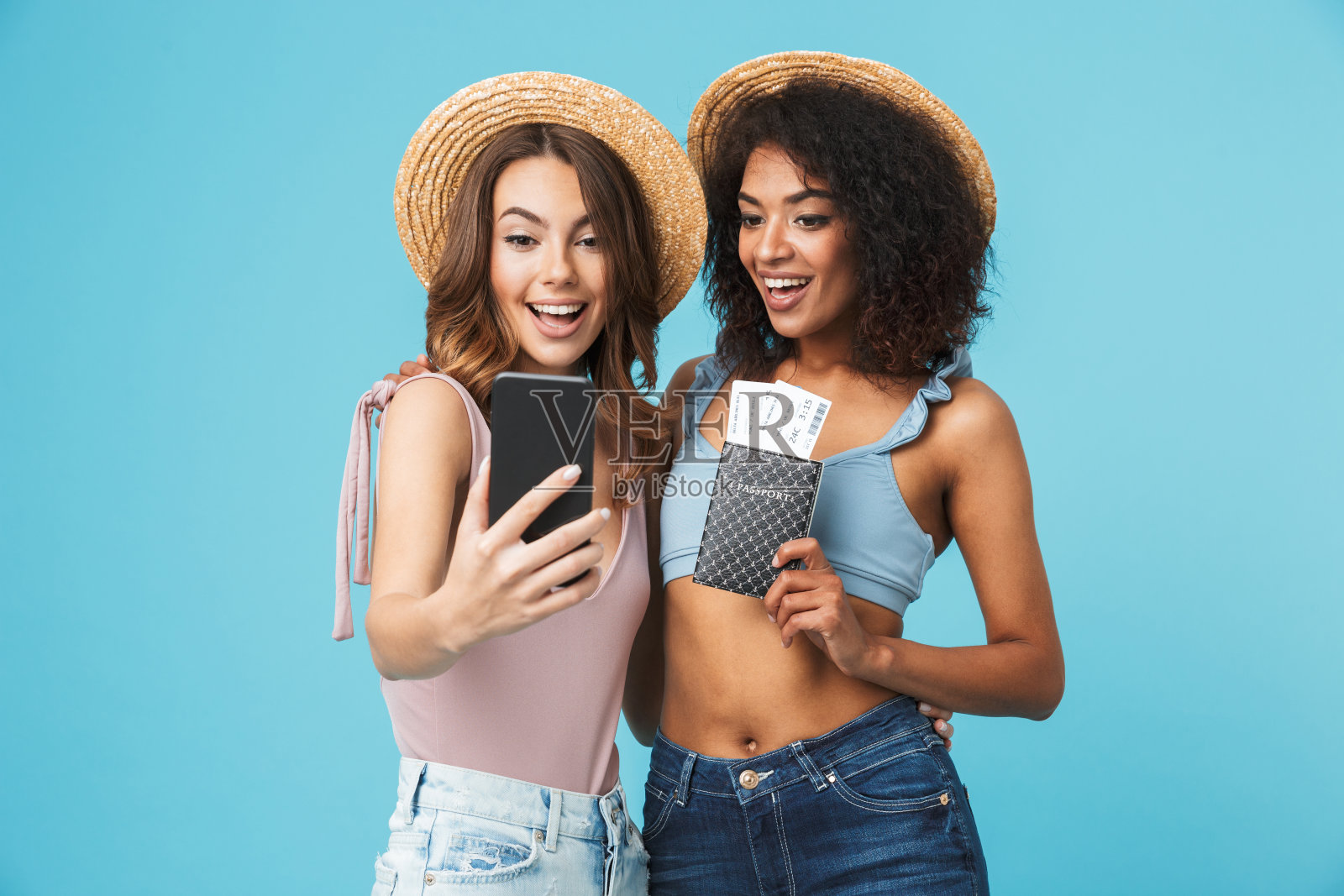 两个多民族夏日女孩拿着护照和机票用智能手机微笑自拍的度假照片，被隔离在蓝色背景下照片摄影图片
