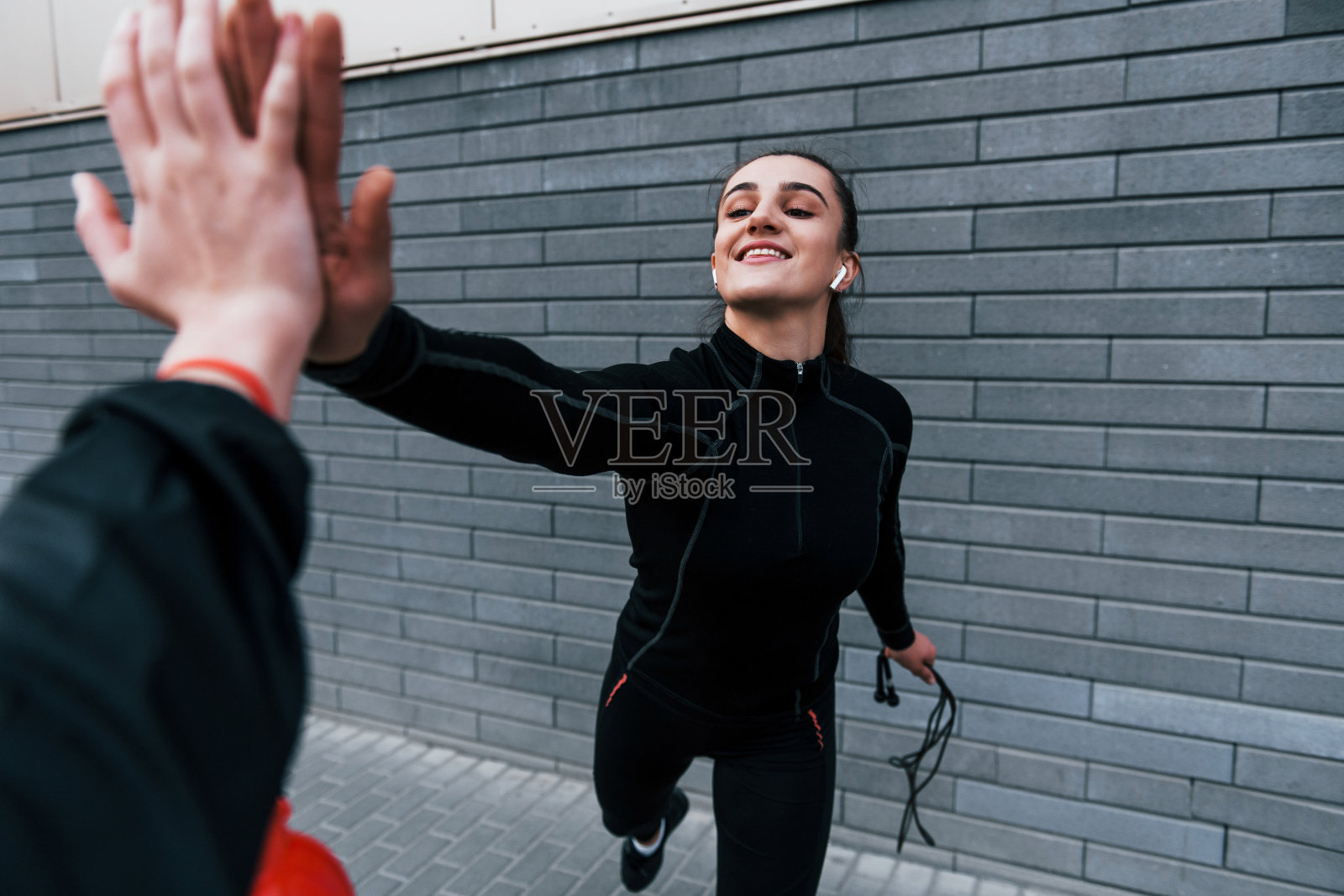 年轻的运动女孩在黑色运动服站在户外与跳绳在灰色墙壁和给击掌照片摄影图片