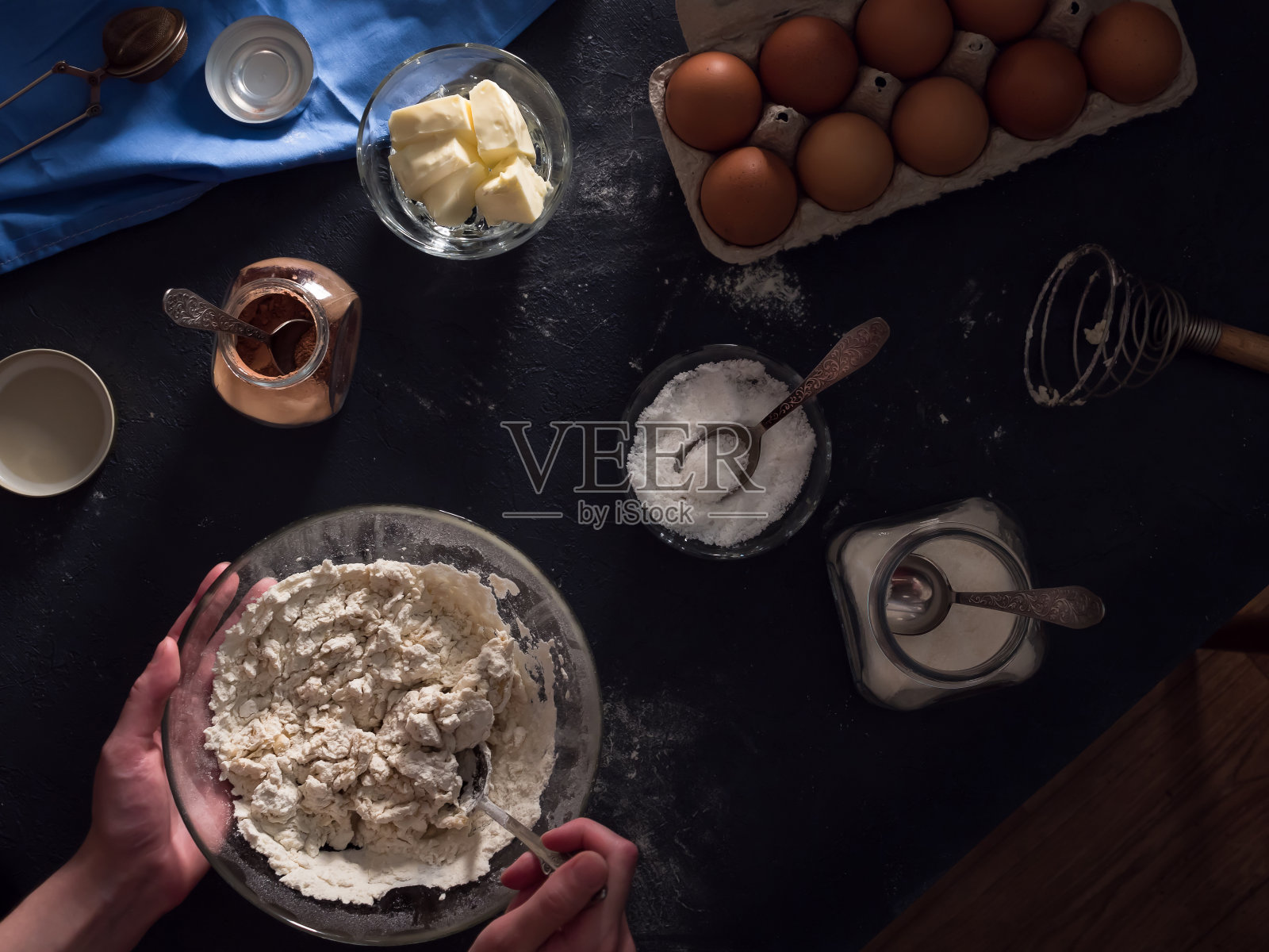 烹饪蛋糕。桌子上放着做馅饼的原料(面粉、鸡蛋、可可、黄油、糖、盐)、厨房用具和纺织品。揉捏面团。手在画框里。桌子和地板的俯视图。平的。暗的背景照片摄影图片