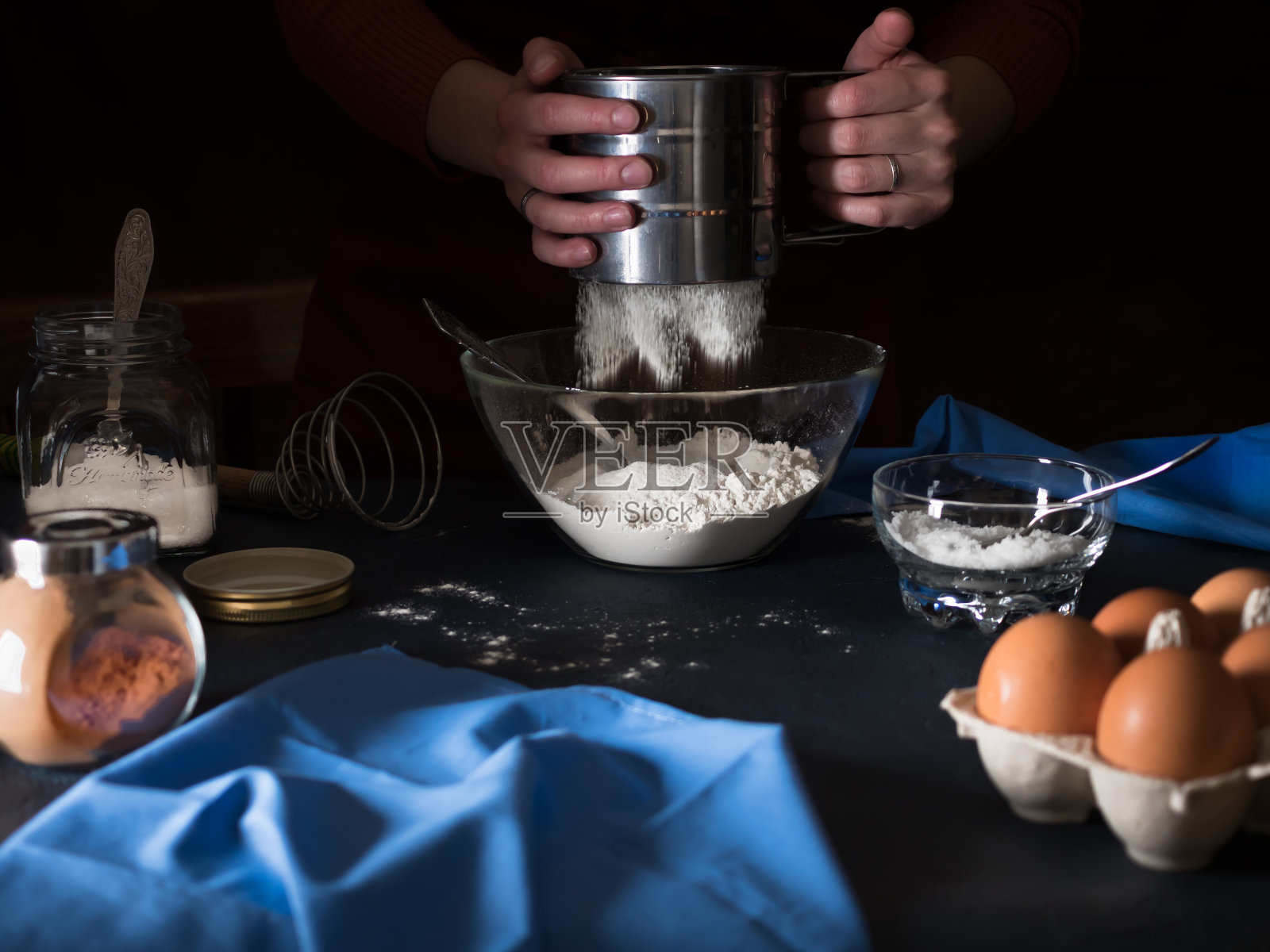 烹饪蛋糕。将面粉筛入碗中。桌子上放着制作馅饼的原料(面粉、黄油、糖、盐、可可、鸡蛋、酵母)、厨房用具和纺织品。背景是一个穿着围裙的女孩。暗的背景照片摄影图片