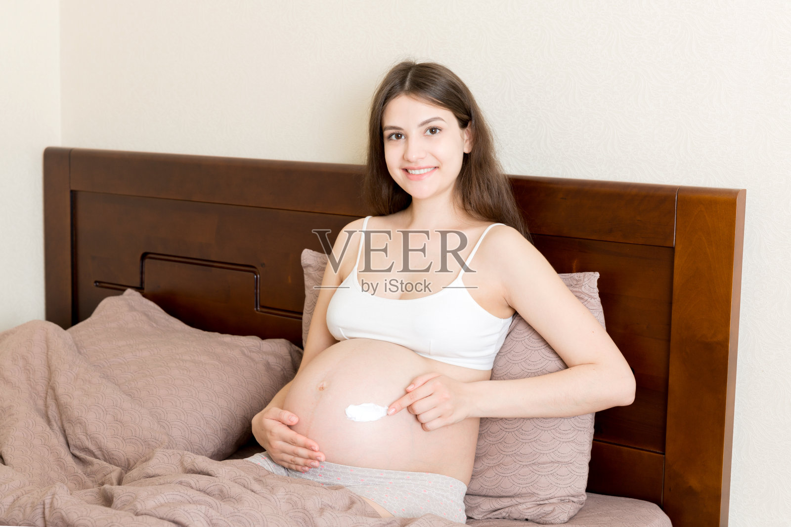 一个怀孕的女孩坐在家里的床上，把抗妊娠纹霜涂在她的肚子上。怀孕、母性、准备和期望的概念照片摄影图片